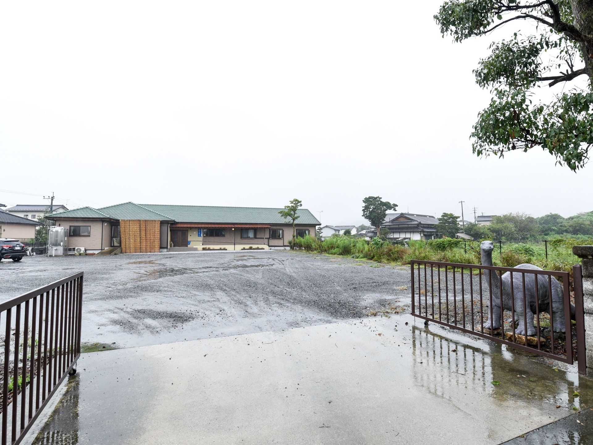 施設敷地の入口は大きな鉄製の両開きの門が設置されている。門の隣には大きな木があり、その下に恐竜の置物がある。