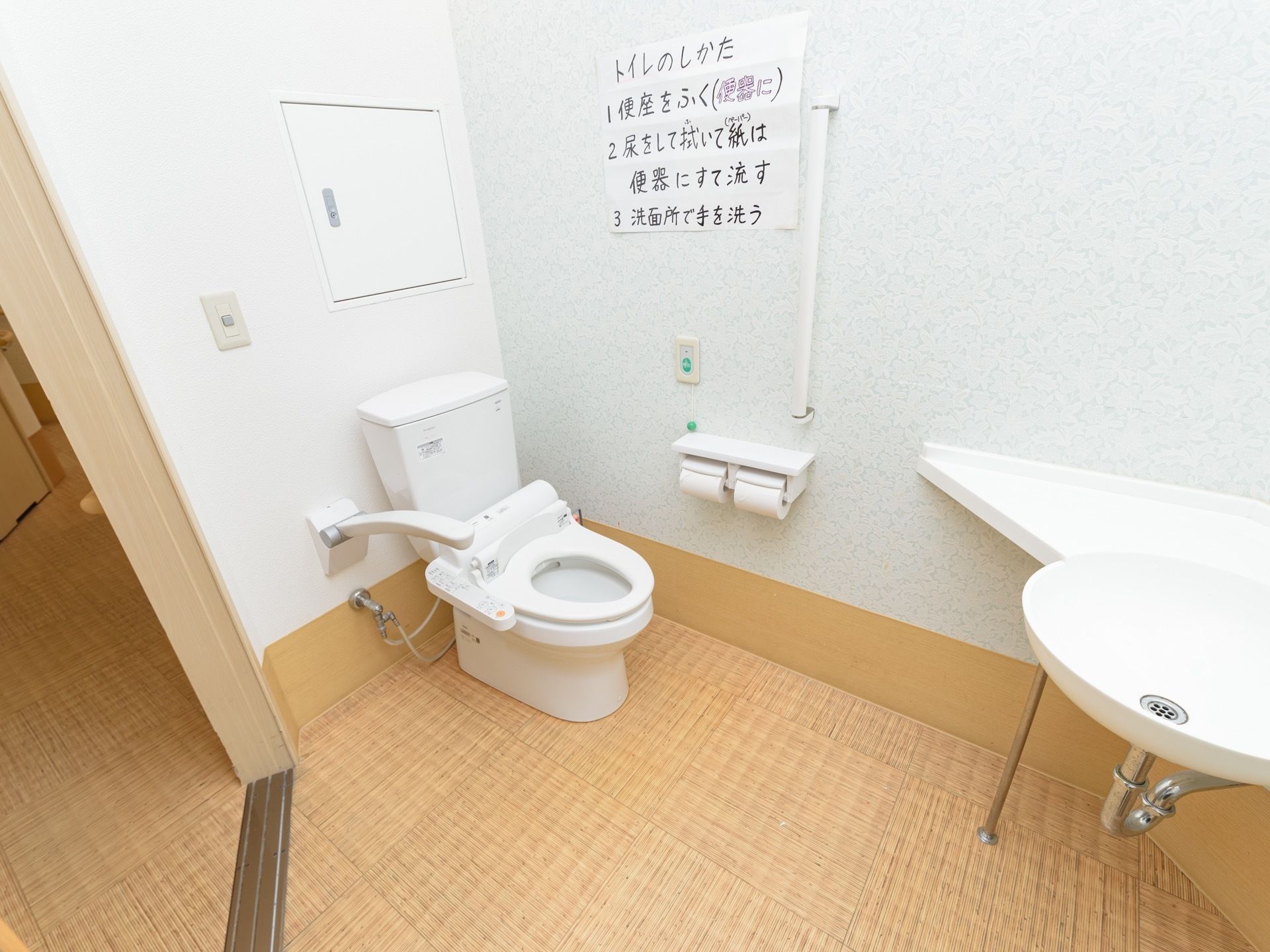 洗面台や手すり、呼び出しボタンが完備された広いトイレである。便器には、温水洗浄便座が設置されている。
