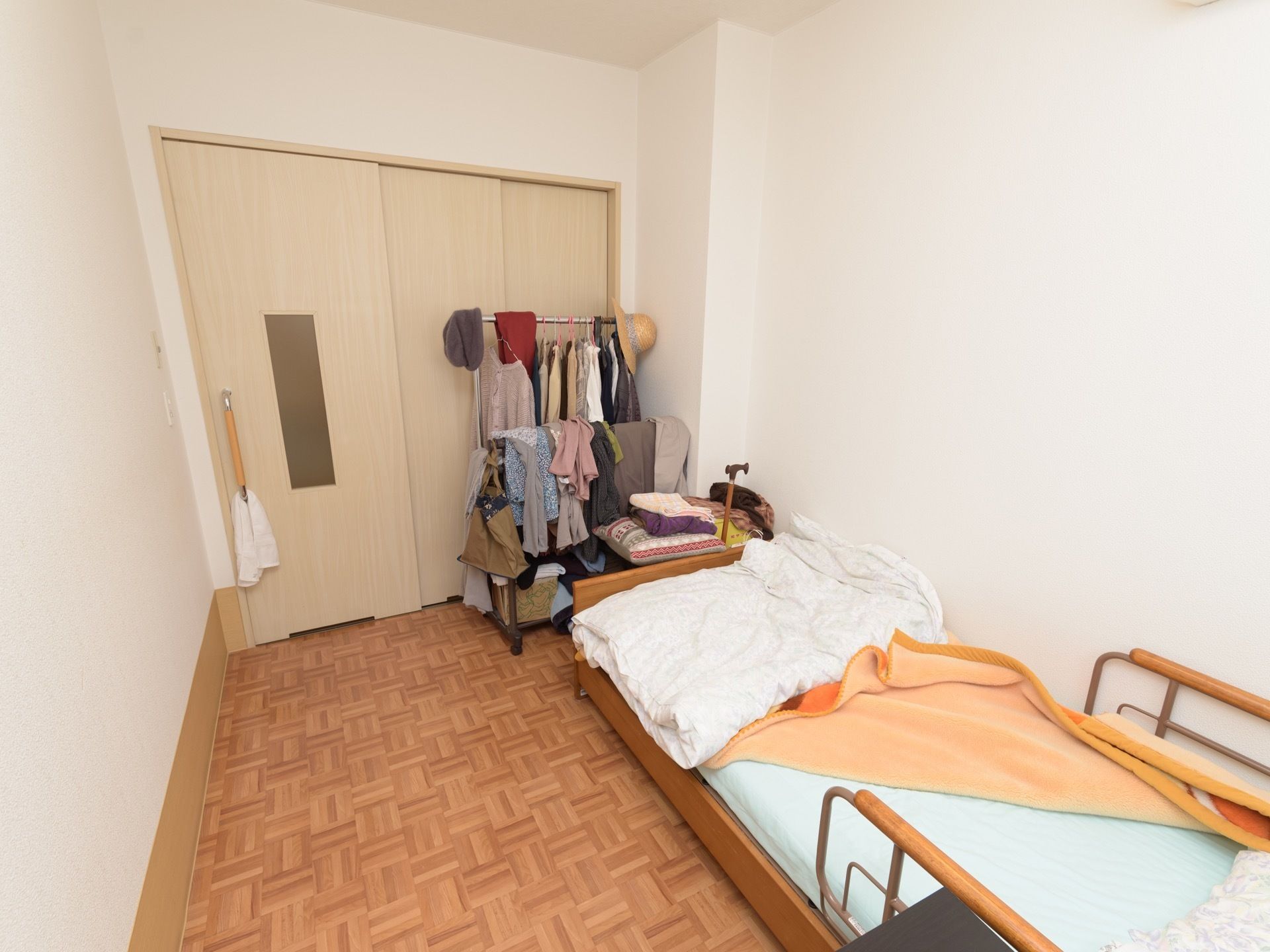 洋室タイプの居室である。壁際に介護ベッドが設置されており、洋服が掛けられたハンガーラックが置かれている。