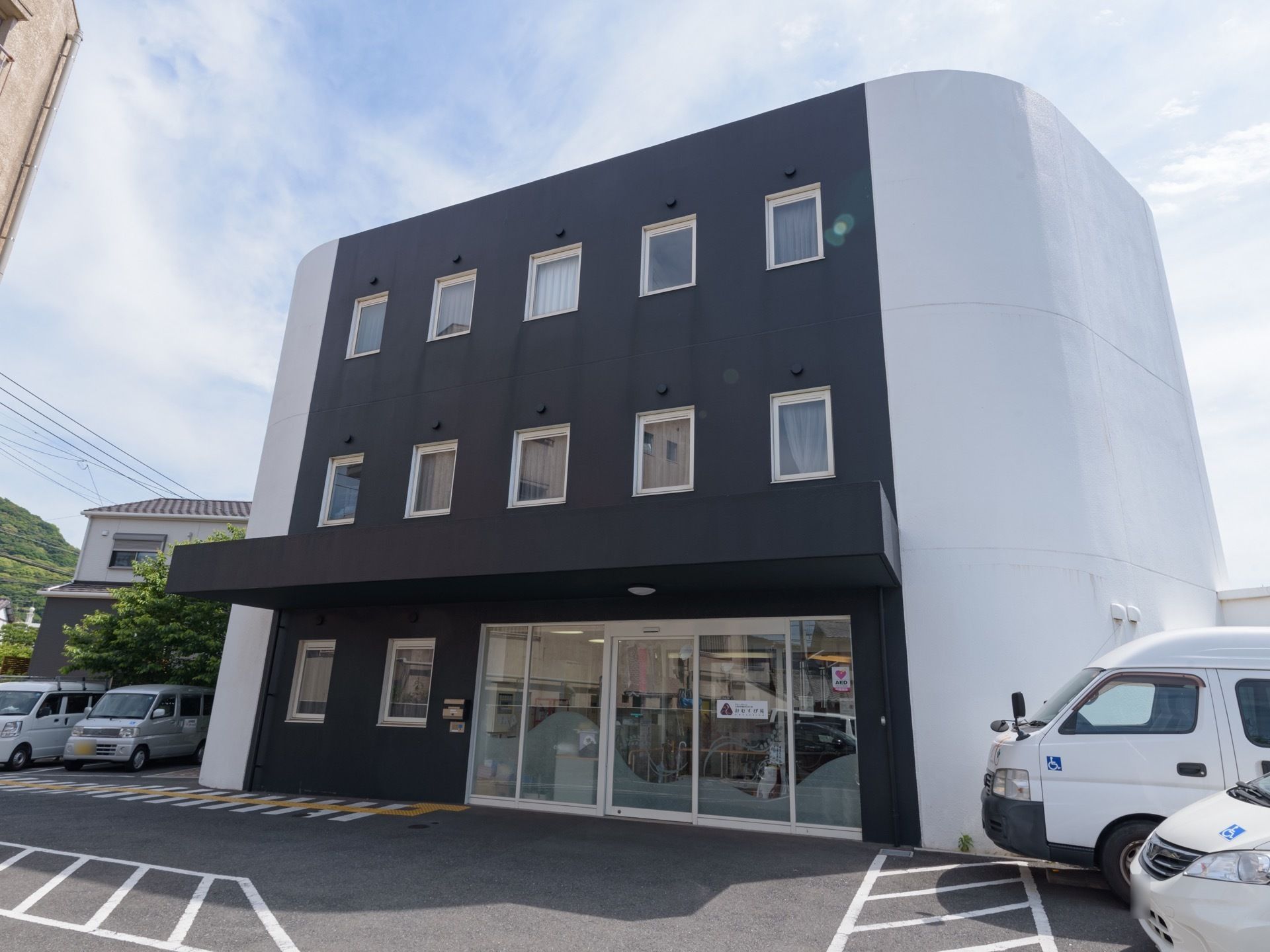施設は白と黒の外壁が特徴的な３階建ての建物である。駐車スペースが建物の両サイドに用意されている。エントランスには、ひさしがついている。
