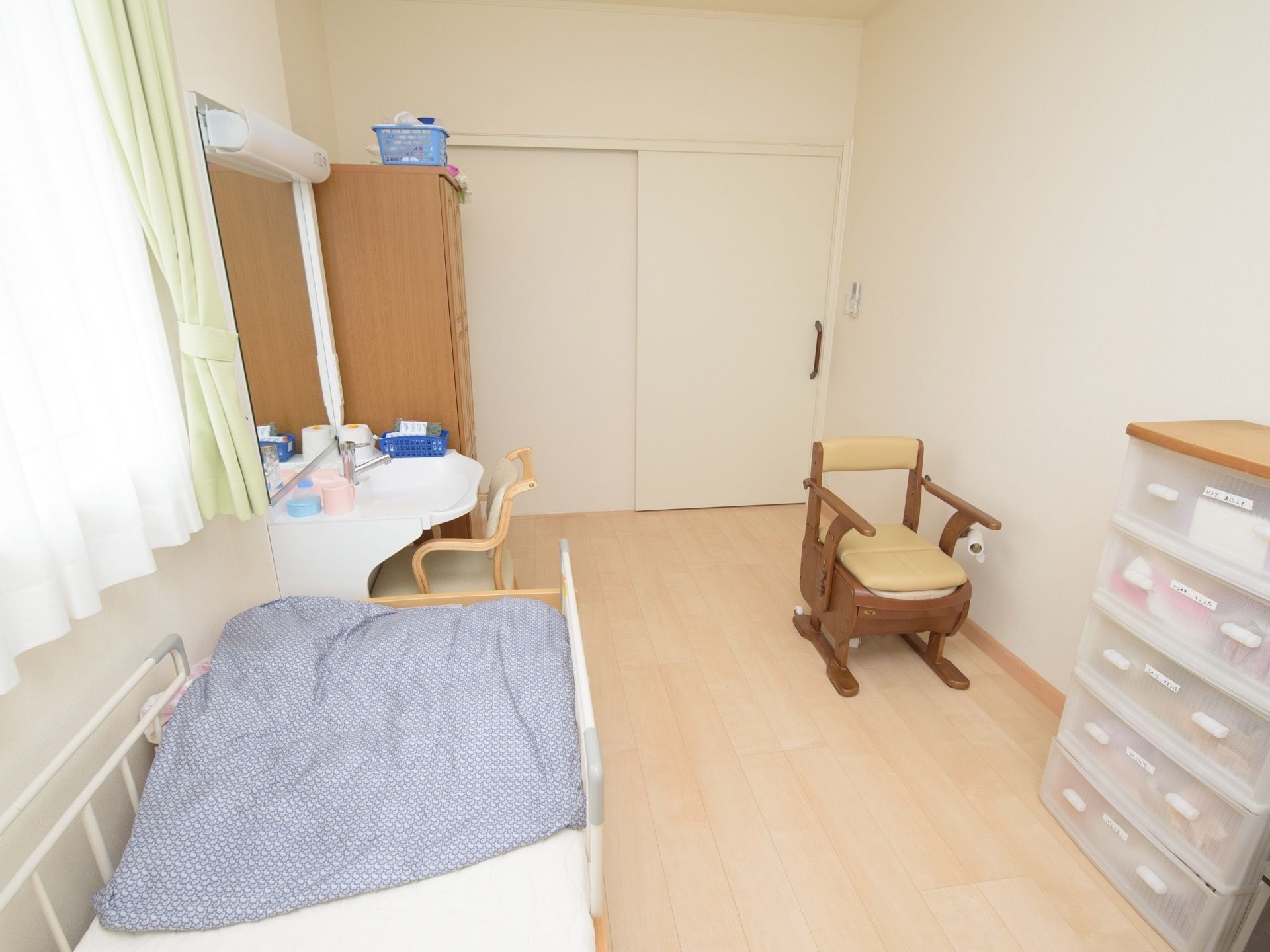 洗面台が備わった居室である。介護ベッドやクローゼットが壁際に設置されている。椅子や家具調のポータブルトイレが置かれている。
