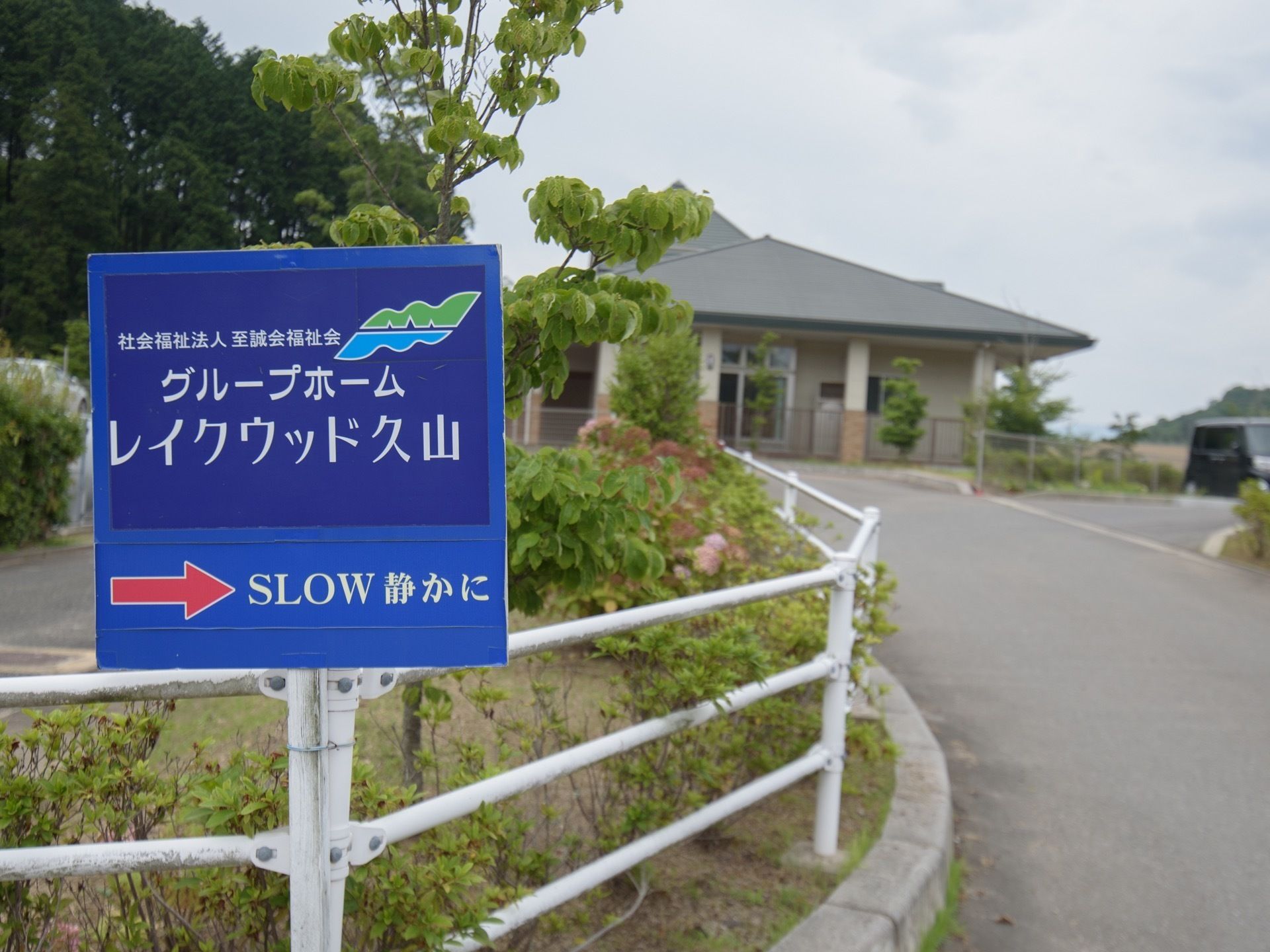 施設の建物前の玄関アプローチには、左側にガードパイプが設置された植え込みがある。施設名が書かれた看板が立てられている。