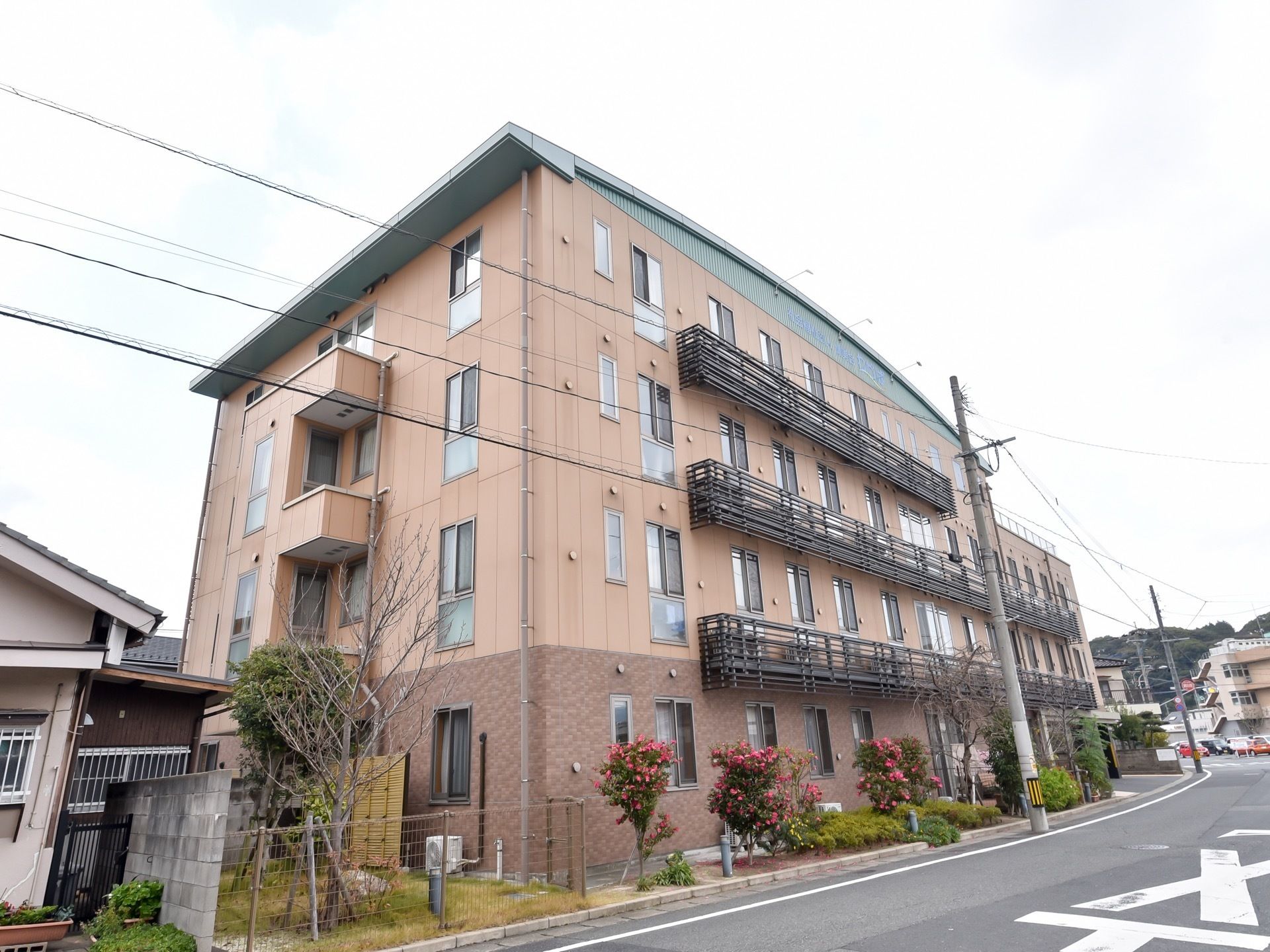 施設の外壁はブラウン色になっており、４階建ての大きな建物である。道路沿いの居室にはベランダが設置されており、室外機が置かれている。