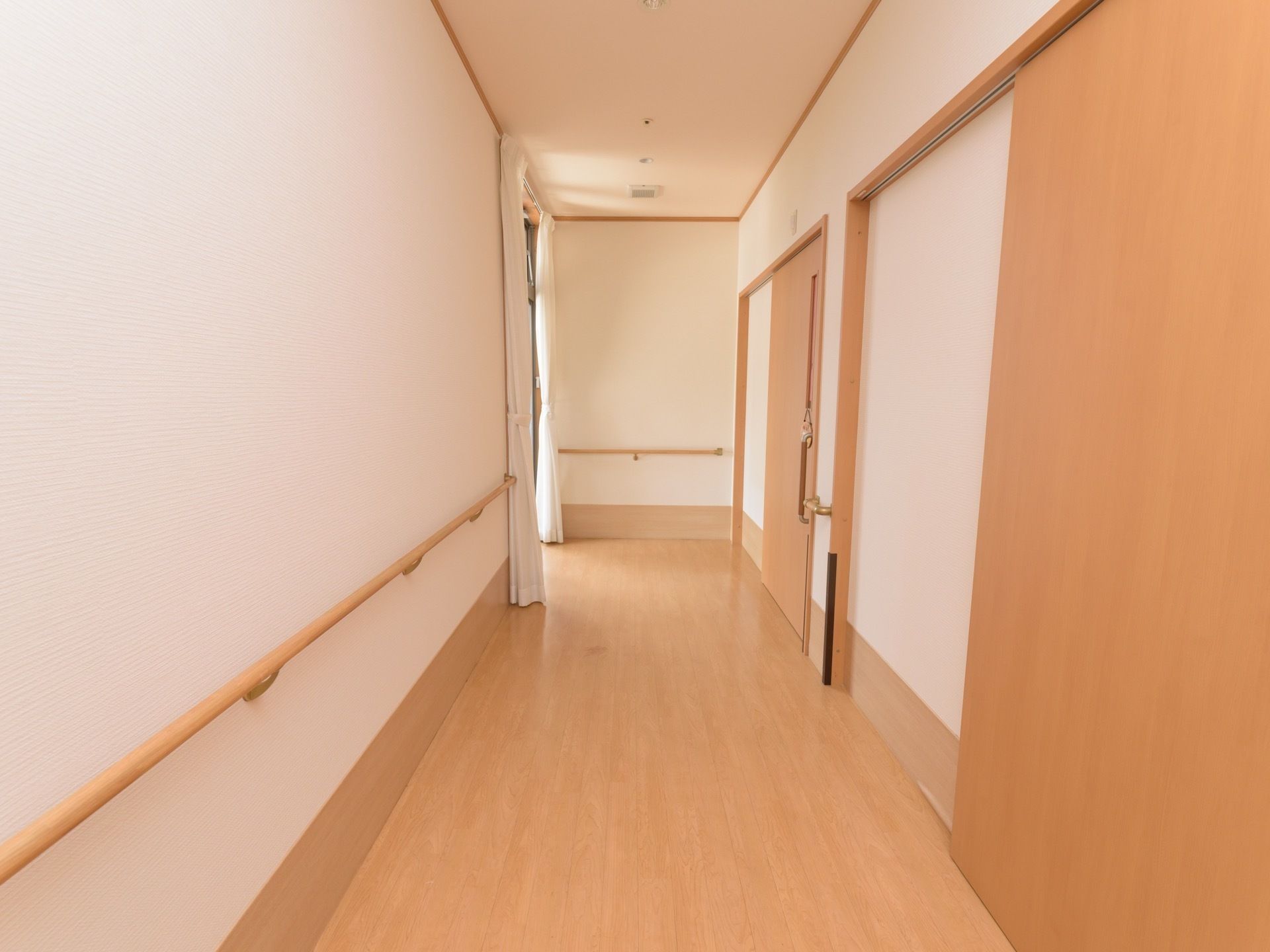 幅広の真っ直ぐな廊下である。手すりが壁に沿って設置されている。右側に居室が設けられており、スライドドアが扉に採用されている。