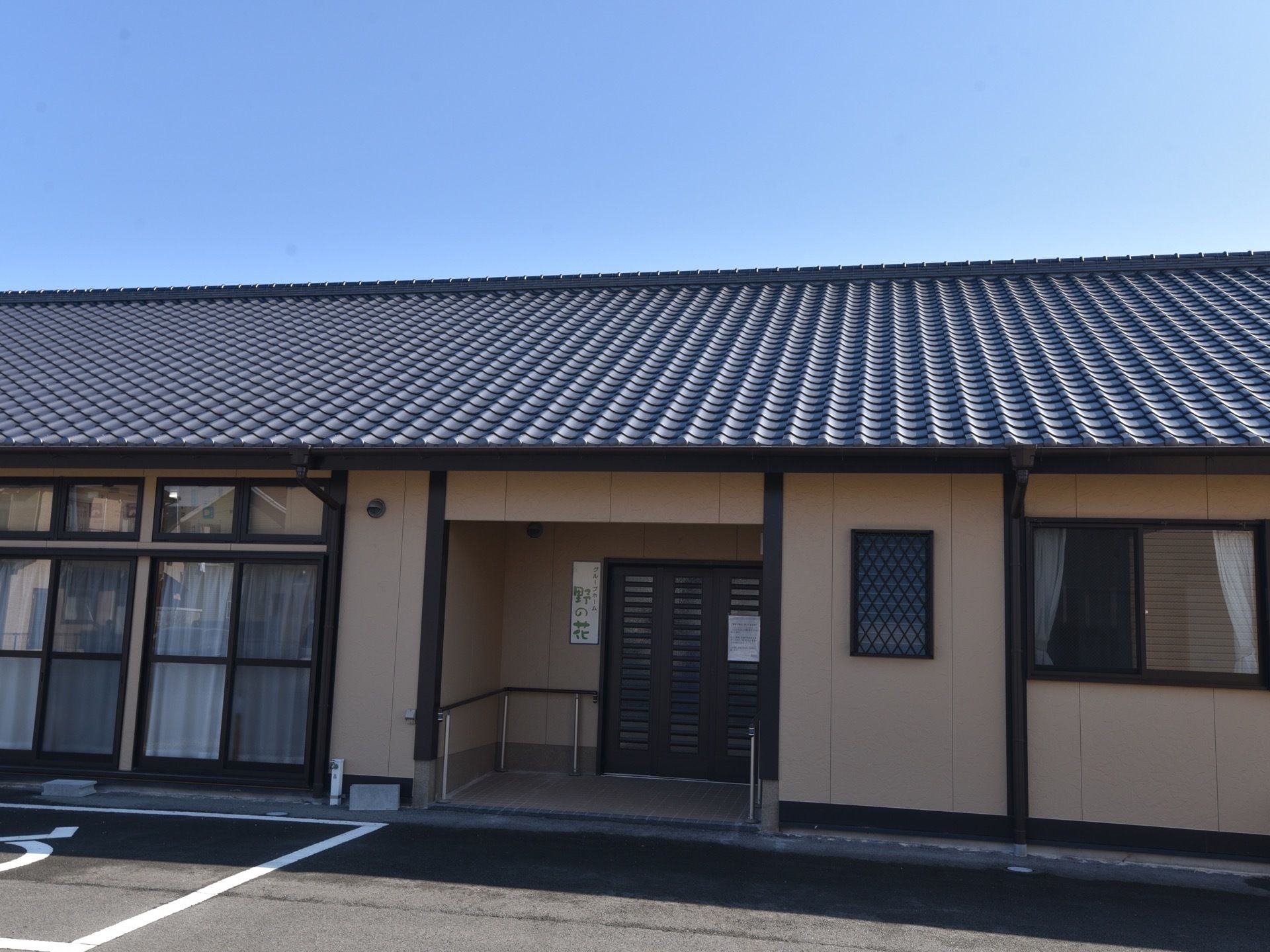 黒い瓦屋根の平屋建ての建物がある。壁は濃いベージュで、窓や柱の縁取り部分が黒に塗られている。玄関には手すりがある。
