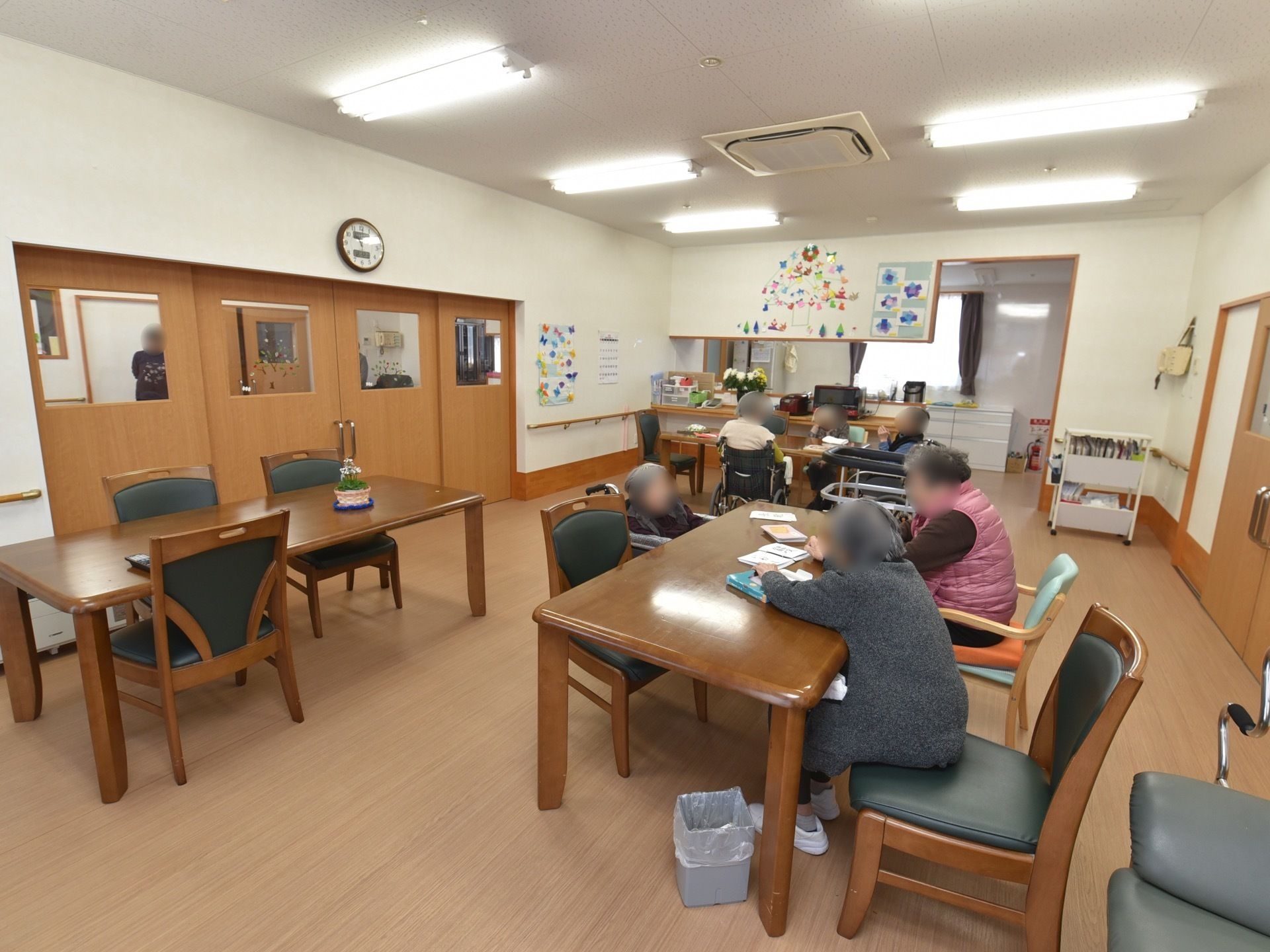 大きな茶色の扉のある広い談話室。テーブルと椅子が置かれている。奥にはキッチンカウンターがあり、壁には飾りつけがされている。