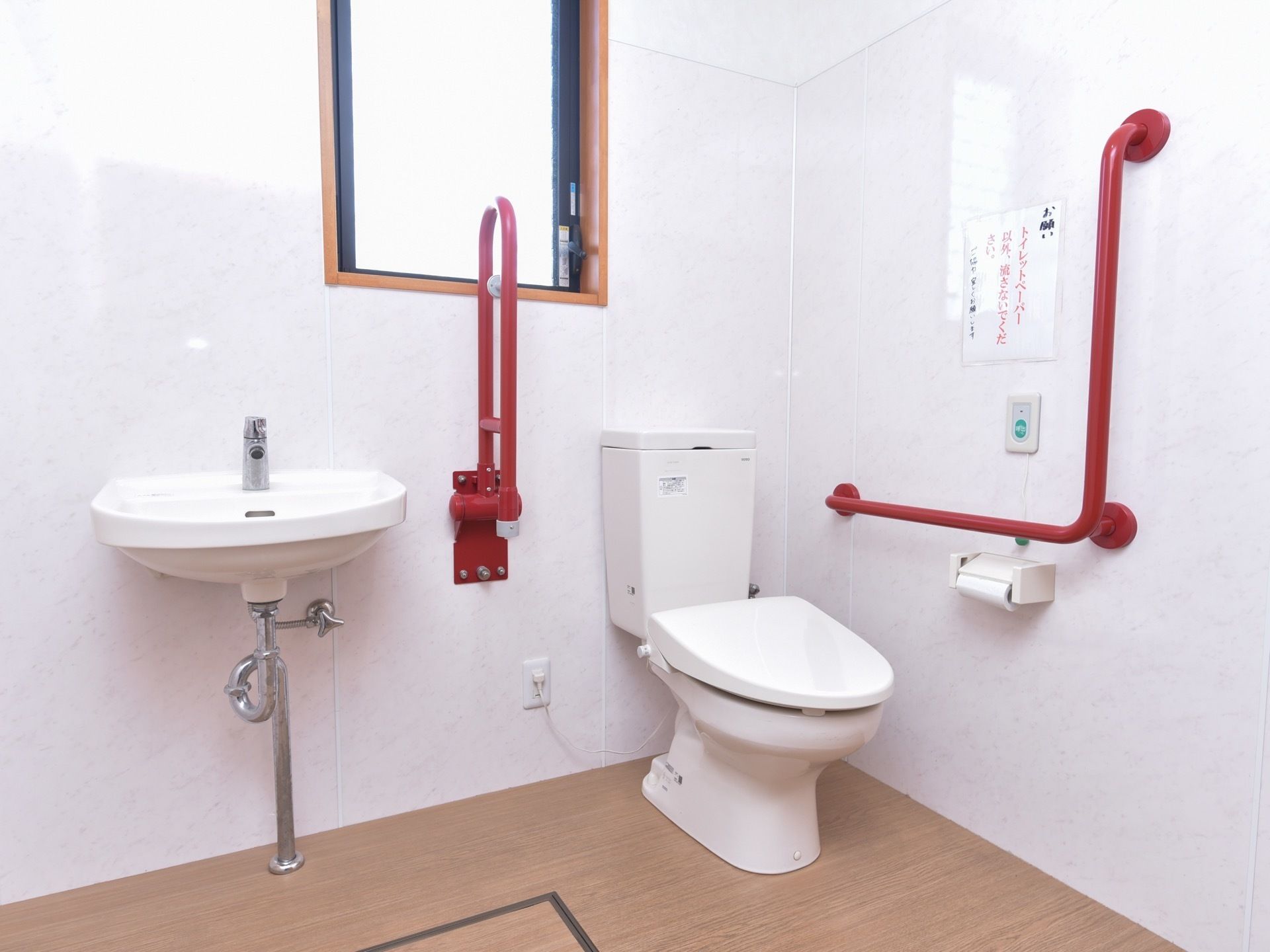 白い壁に囲まれた広いトイレがある。縦長の窓があり、洗面台がある。壁には目立つように赤い手すりが設置されている。