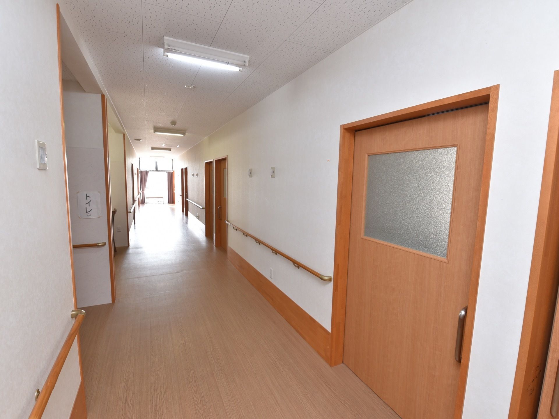 白い壁とブラウンの扉が並んでいる廊下がある。扉にはすりガラスの窓がはまっている。突き当りは窓があり、外光が差し込んでいる。