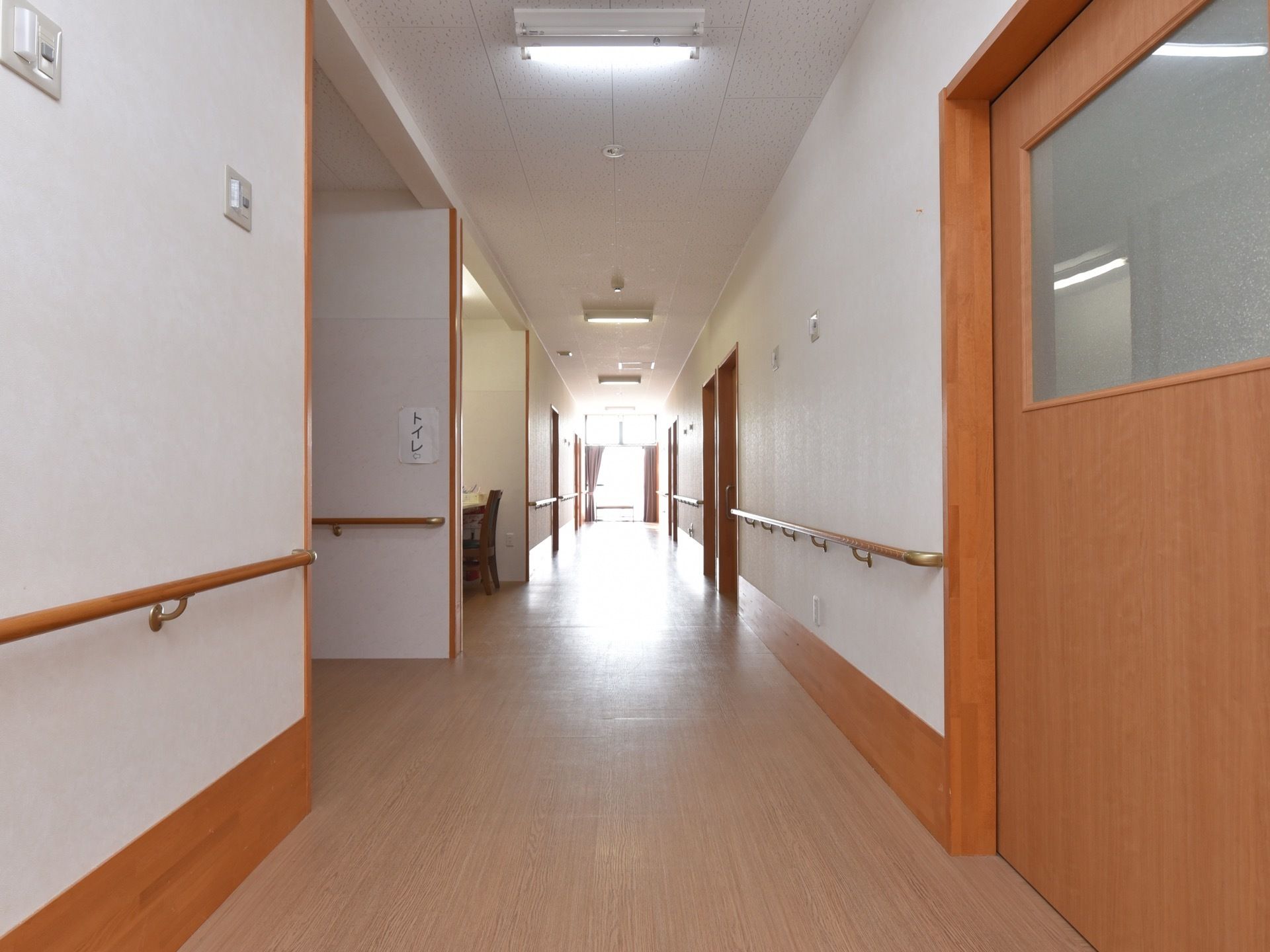 入居者様の居室のドアが並んでいる長い廊下がある。壁は白く、床と扉はライトブラウンになっていて統一感のあるデザイン。壁には手すりがある。