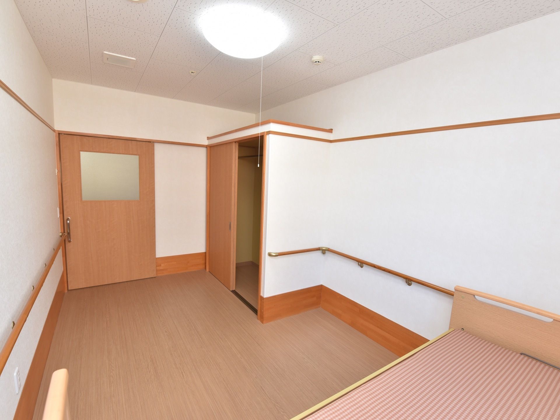 白い壁とライトブラウンの床と扉のある居室。窓際にはベッドが置かれており、大きなクローゼットがある。壁には手すりが設置されている。
