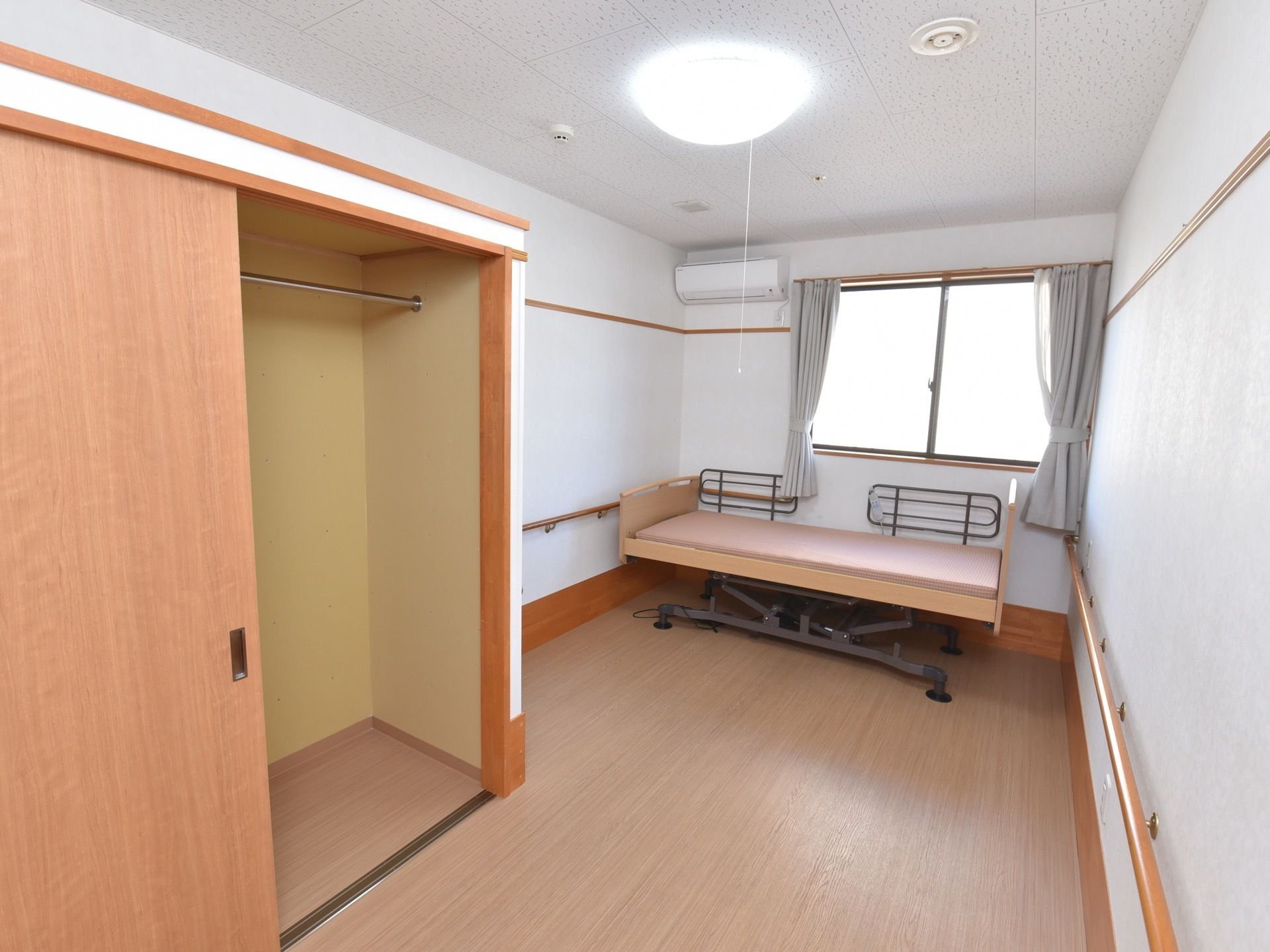 白い壁に手すりが設置されている入居者様の居室。窓の側には介護用ベッドが置かれ、側面に柵がついている。大きなクローゼットがある。