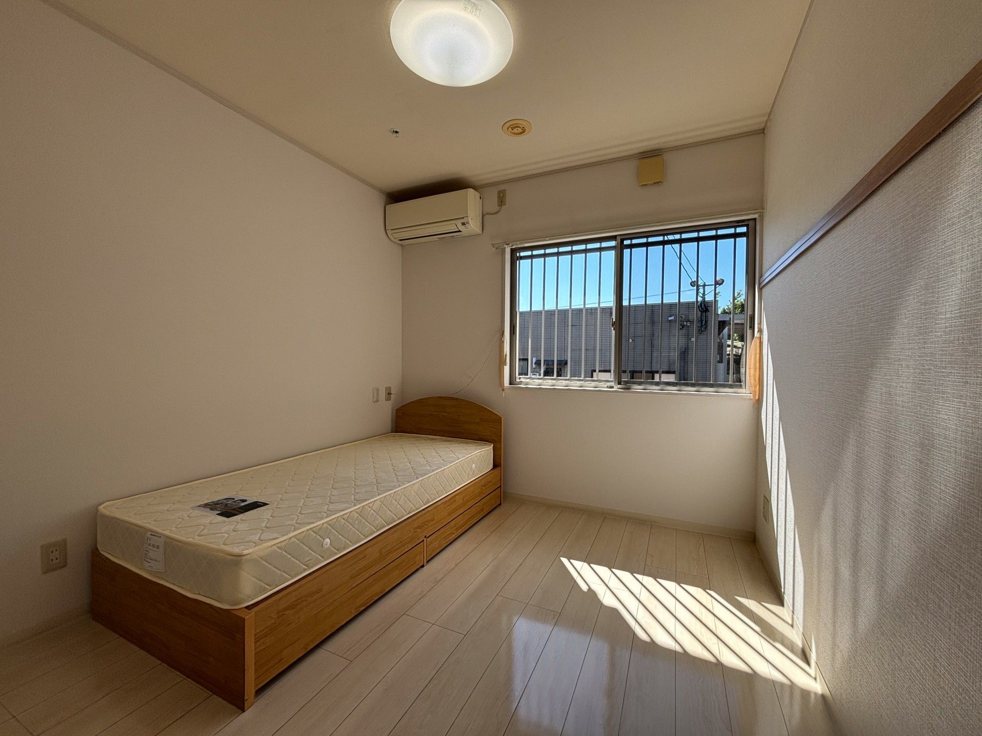 エアコンやベッドが設置されているフローリングの部屋にはレースカーテンとカーテンが付いた窓があり明るい雰囲気。
