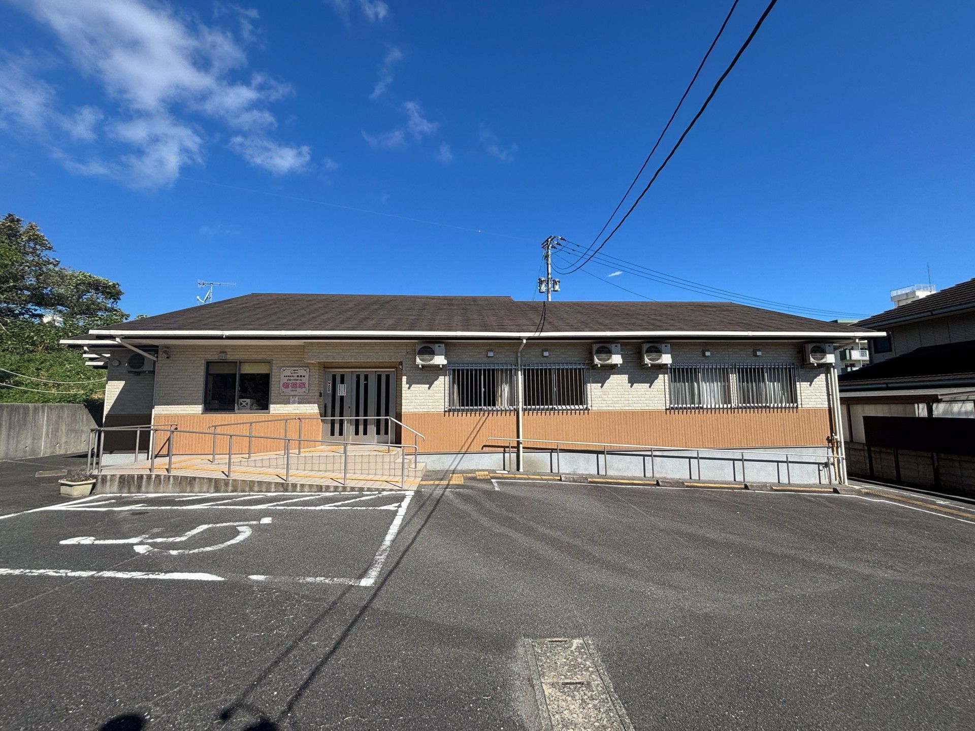 ベージュと山吹色の外壁の平屋建ての建物にはグレーの屋根が付き、玄関先には手すり付きのスロープがある。