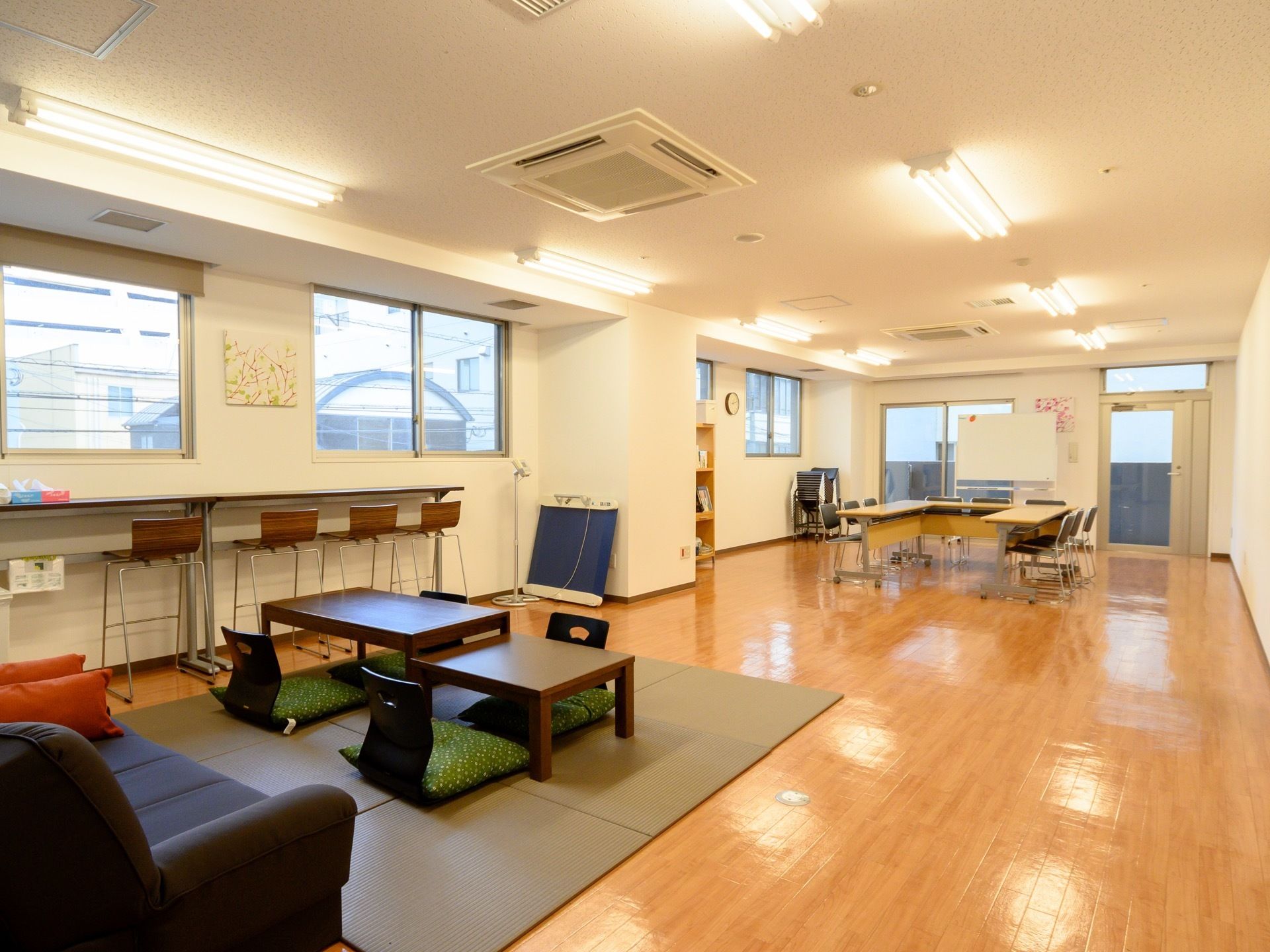 奥に移動式の長机と椅子がある。手前には壁際にカウンターと椅子、敷物の上には座椅子、その前にソファーがある。