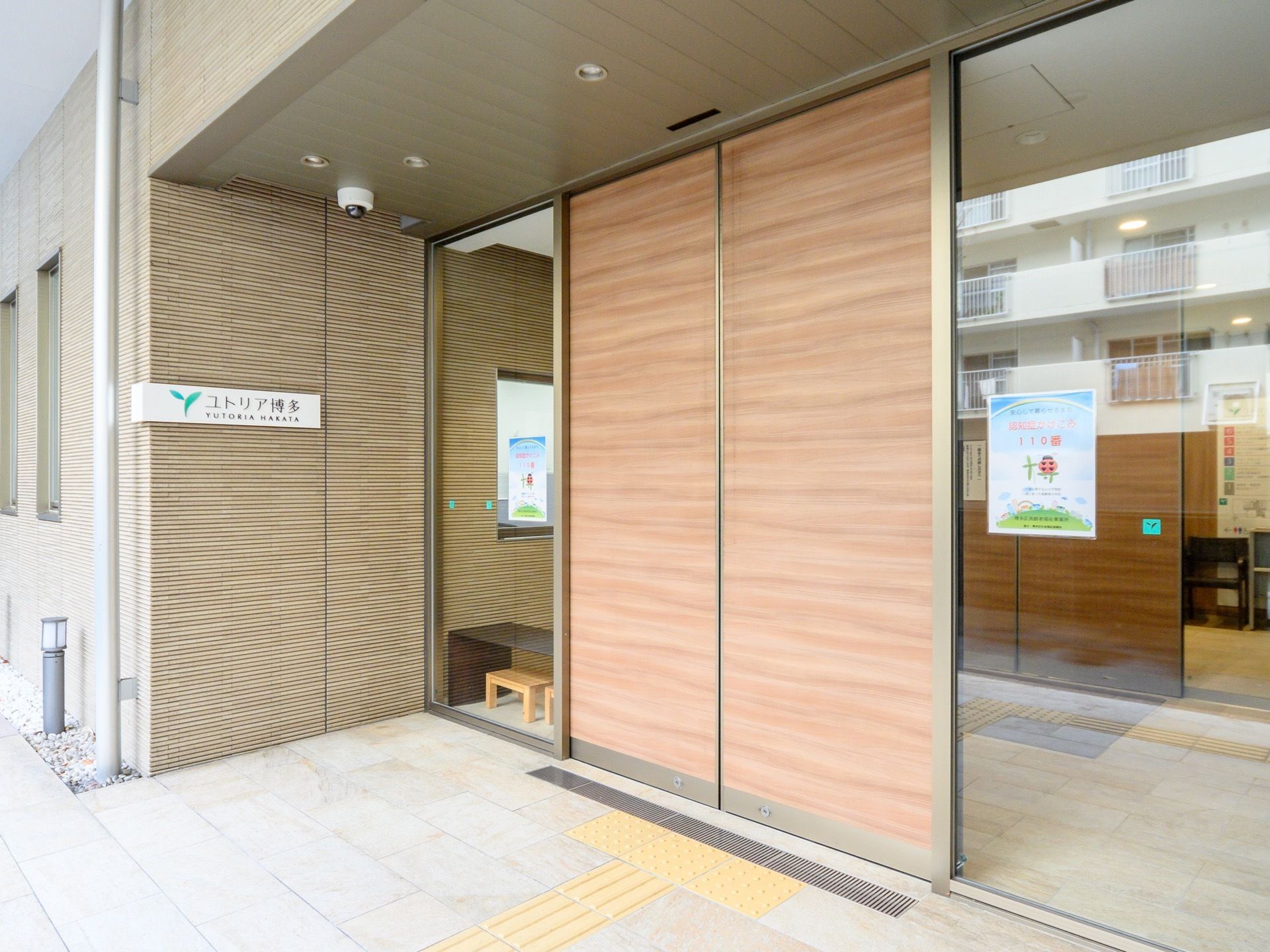 ガラス張りの壁の真ん中に茶色の自動ドアがある。ガラスの部分にはチラシが貼ってある。ドアの奥にはもう１枚自動ドアがある。