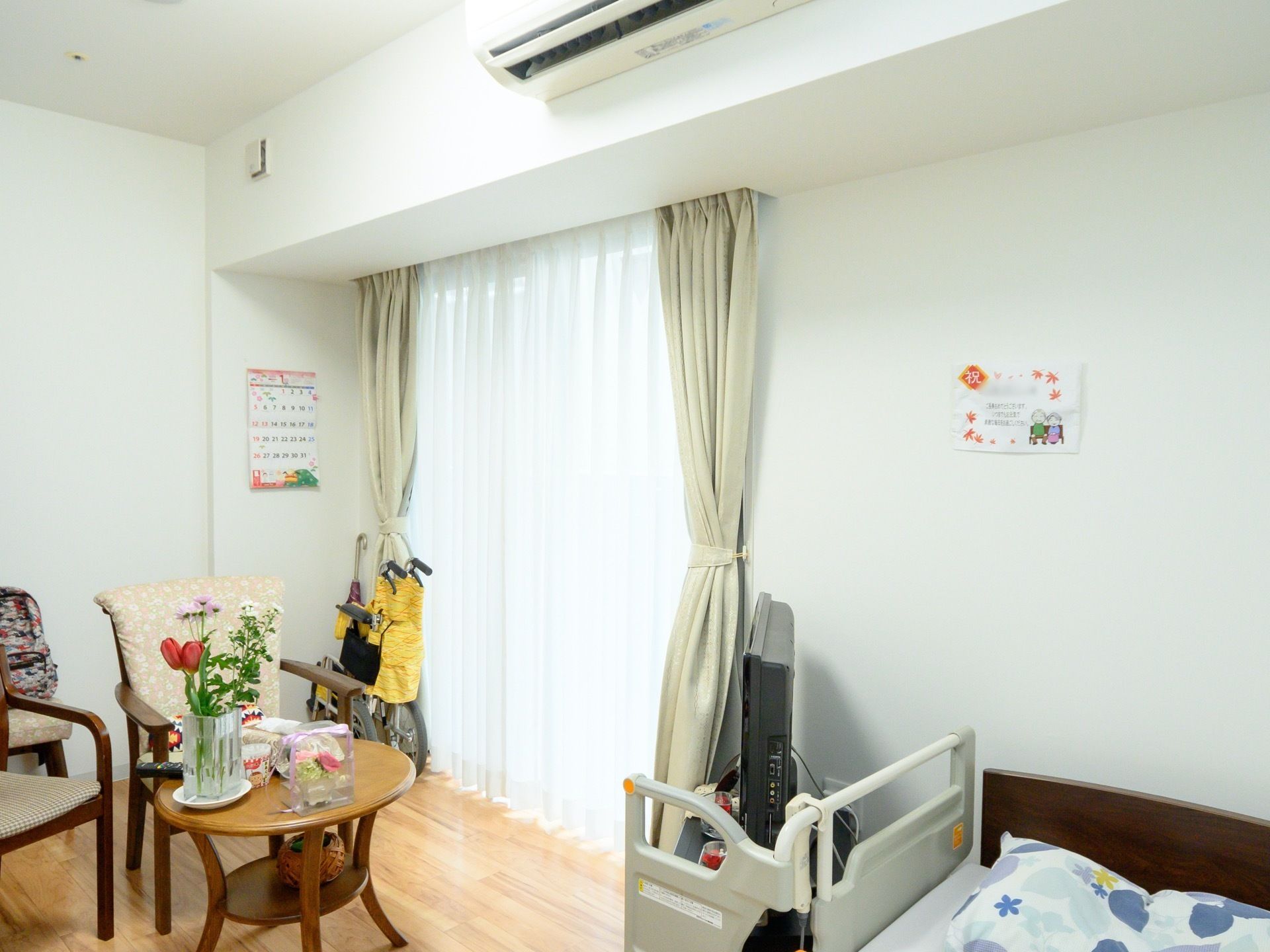 ベランダの窓にはカーテンが２枚掛かっている。肘掛付きの椅子が３脚と丸い机、テレビと介護用ベッドが置かれている。