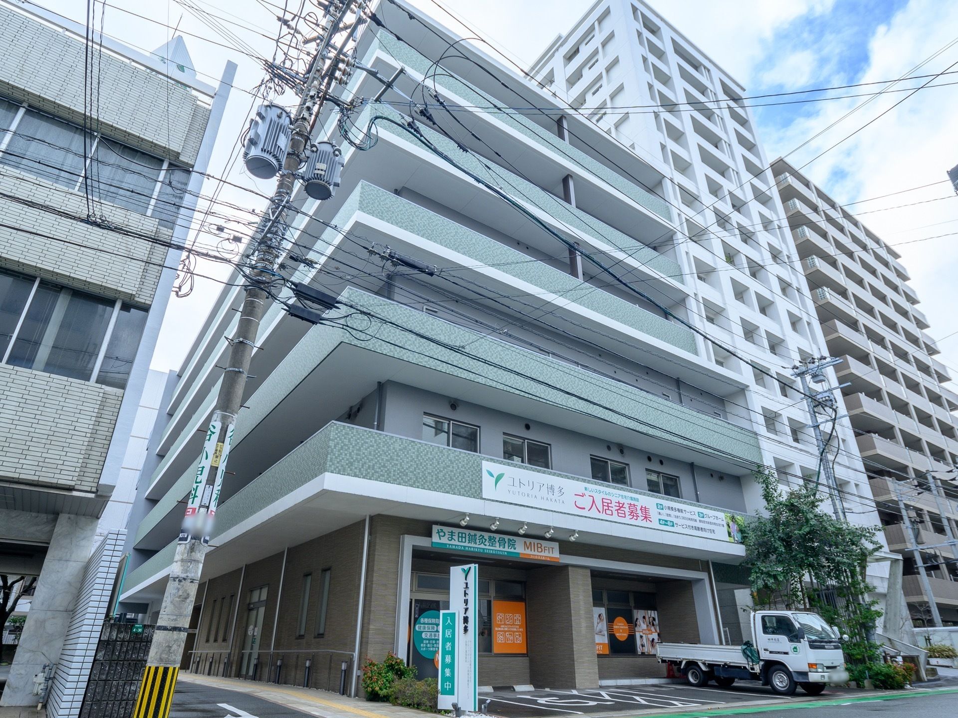 ６階建ての建物。壁は薄い緑と白で１階部分だけベージュ。手前にトラックが停まっている。入居者募集の看板が掛けられている。