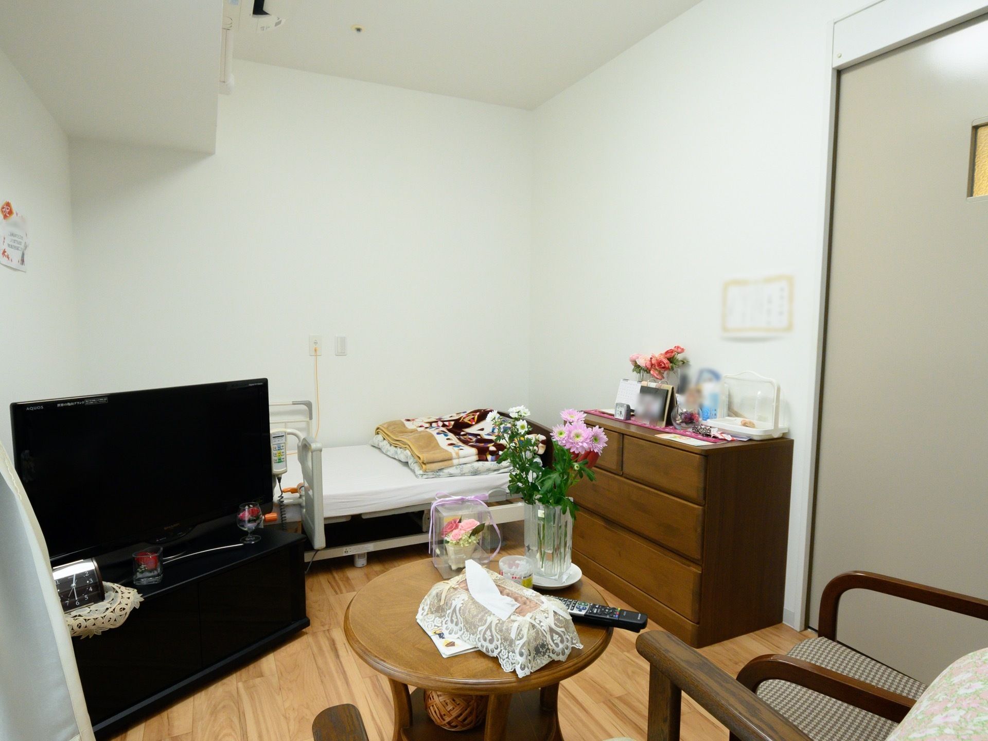 白い壁の居室にテレビとベッド、その隣にタンスが置かれている。丸い机の上に花とティッシュ箱とリモコンがある。