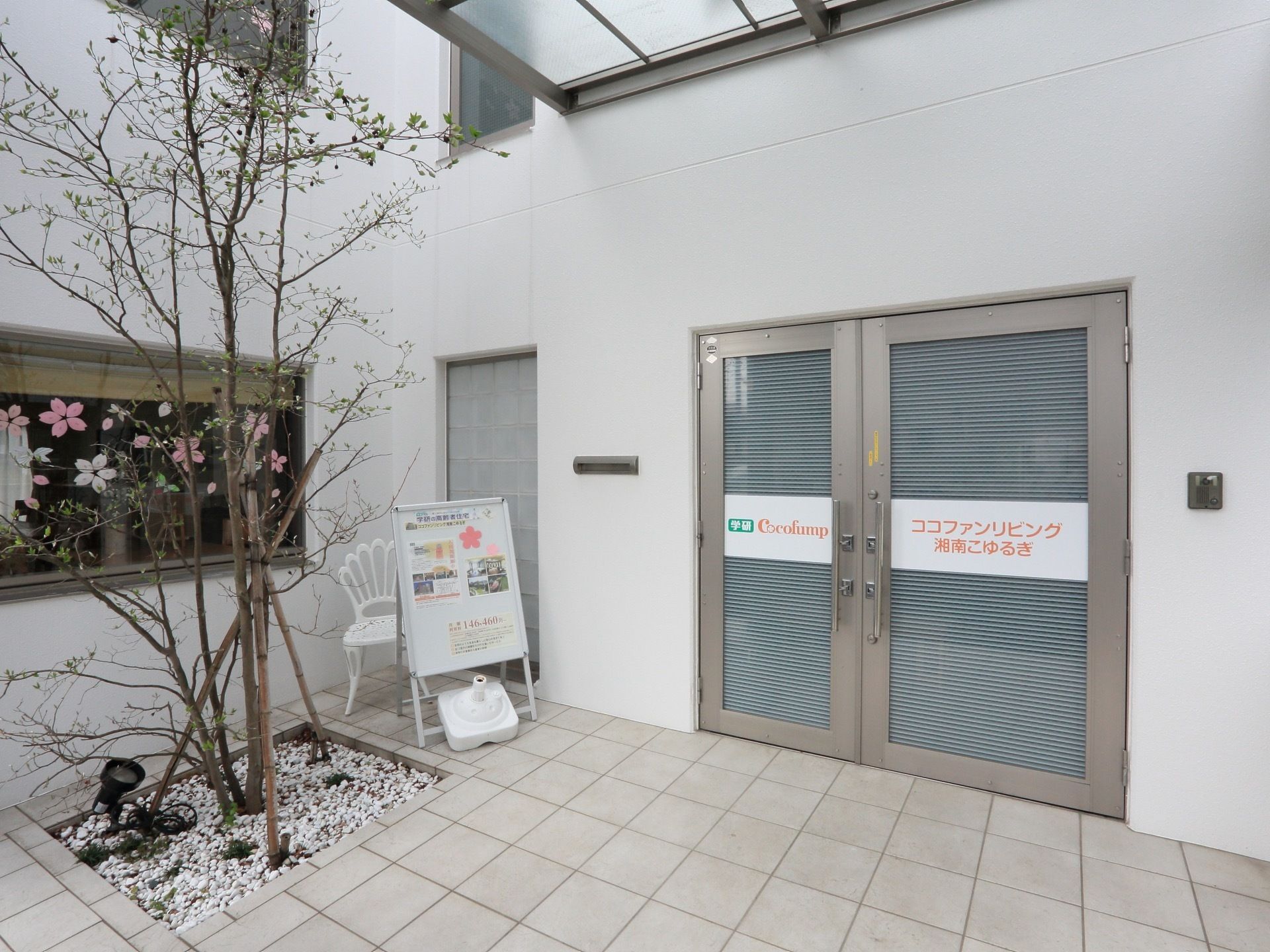 テラスの様な白いタイル張りの玄関である。玄関横には花が咲く木が植えられている。ドアに施設名が書かれている。