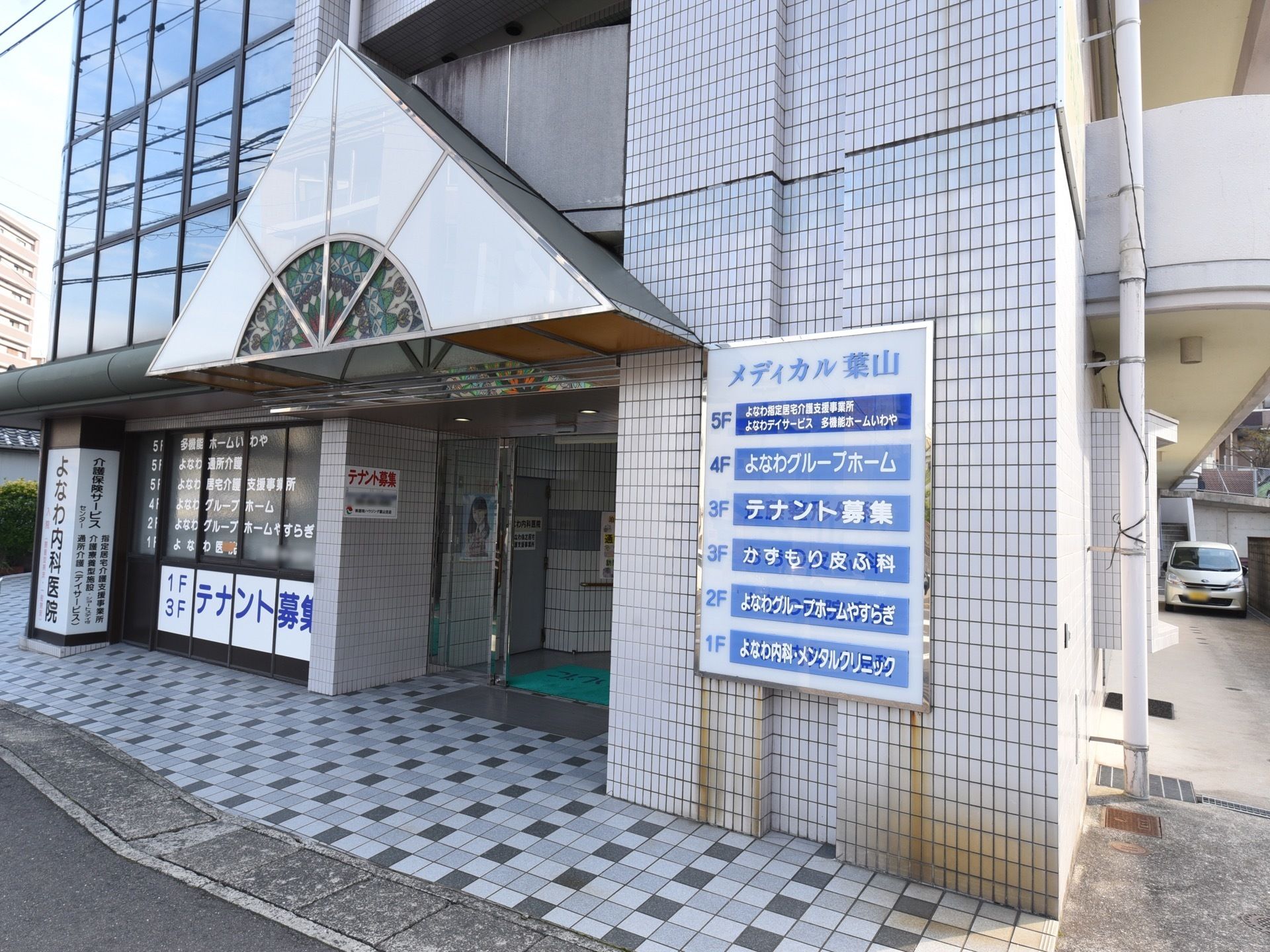 同じ建物内に内科や皮膚科が入居している。外壁にテナントが表示されている。地面はタイルが敷かれている。