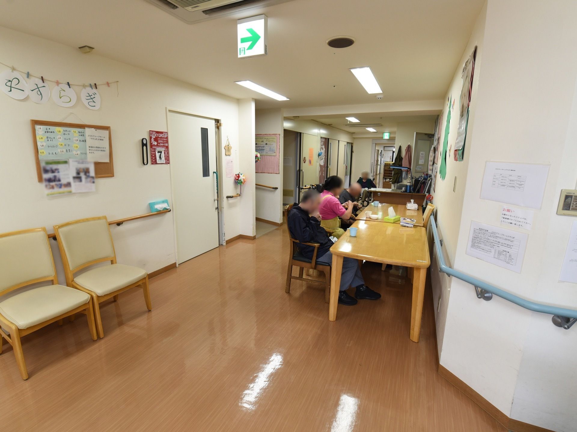 廊下の壁際に１人用の椅子が並んでいる。反対の壁際にはテーブルセットが置かれ、箱ティシュがある。入居者様がくつろいでいる。