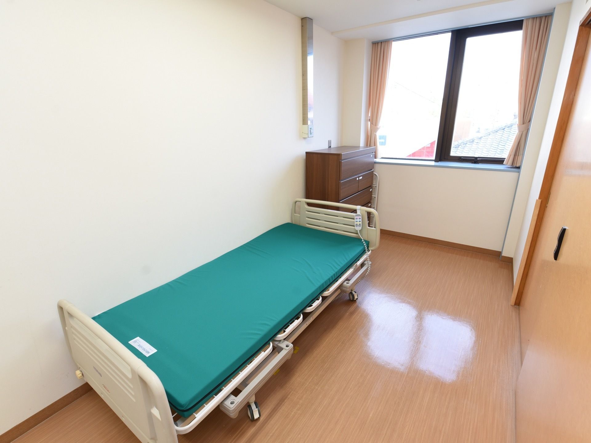 居室には大きな窓があり、ベージュのカーテンがかかっている。壁際に介護用のベッドがあり、その奥にチェストが用意されている。