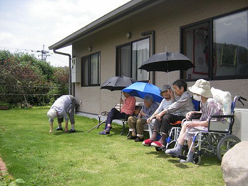 玄関の横にある芝生スペースにはベンチがあり、傘を差した入居者が座っている。草取りをしている人もいる。