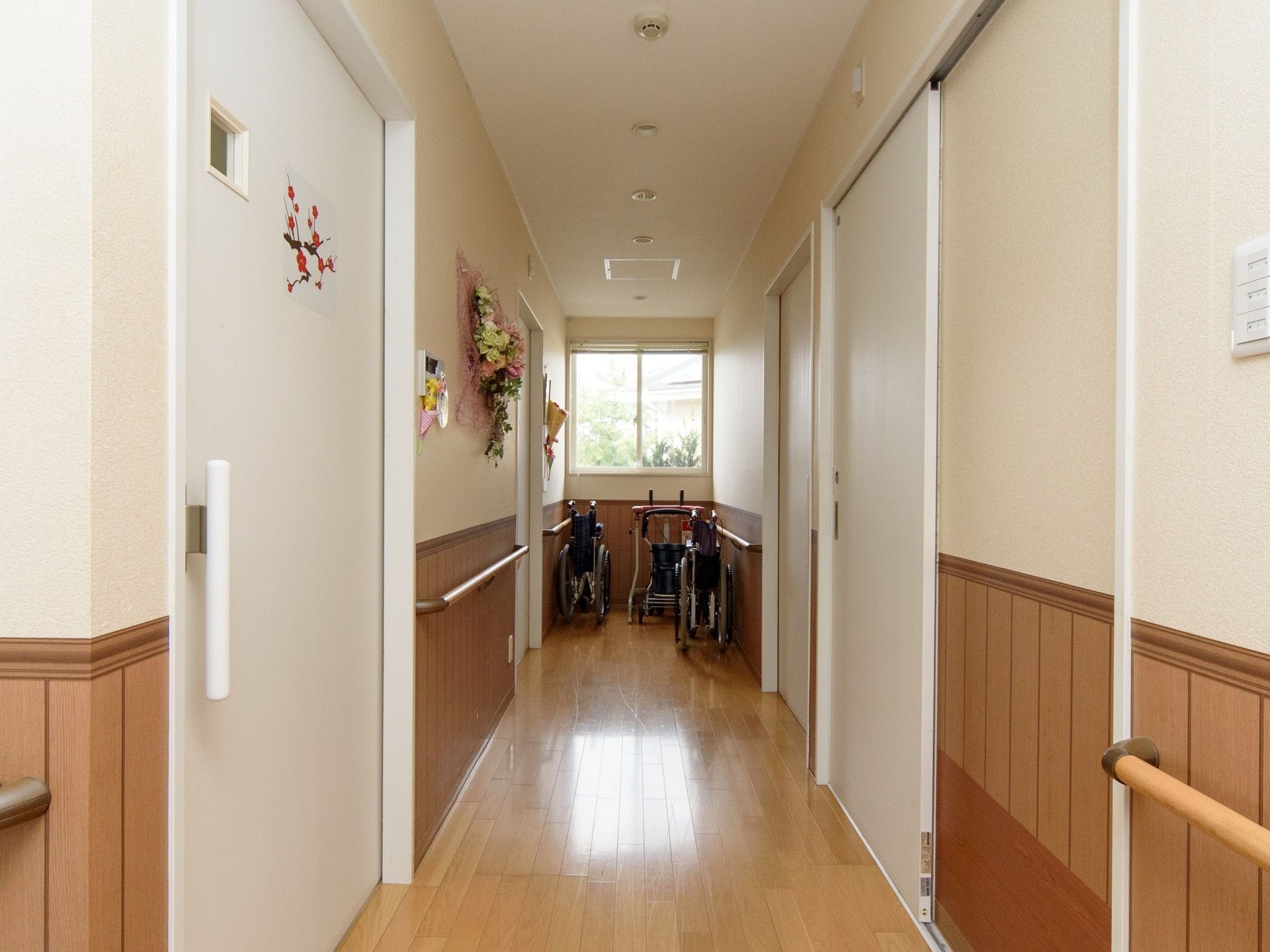 床、壁紙および手すりを木目調で揃えており、落ち着いた雰囲気である。突き当りに介護用品が用意されている。壁に花が飾られている。