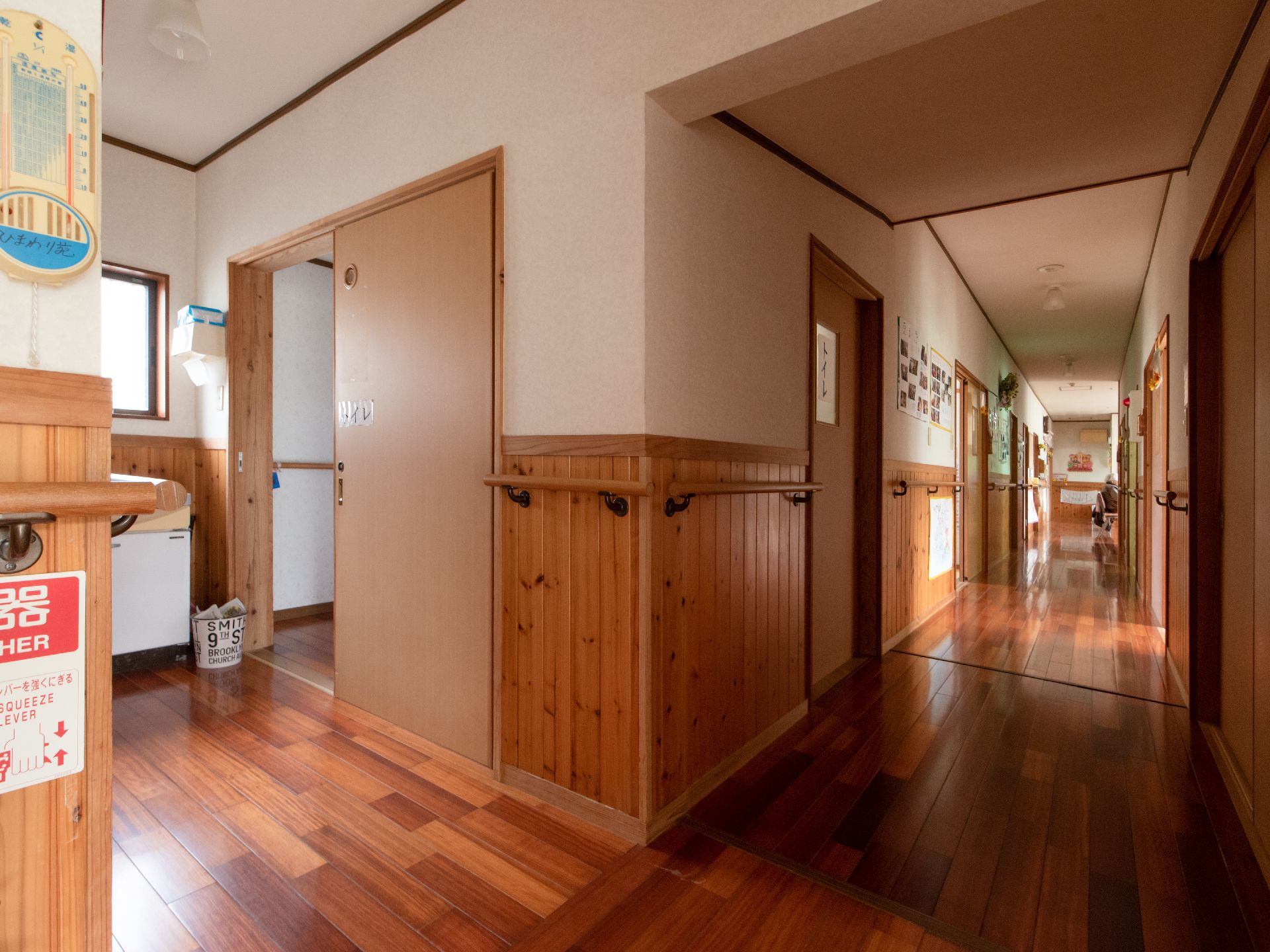 壁が半分板張りになった廊下で、左手前にトイレへの扉がある。右奥へ廊下が伸び、両側に居室への扉が並んでいる。