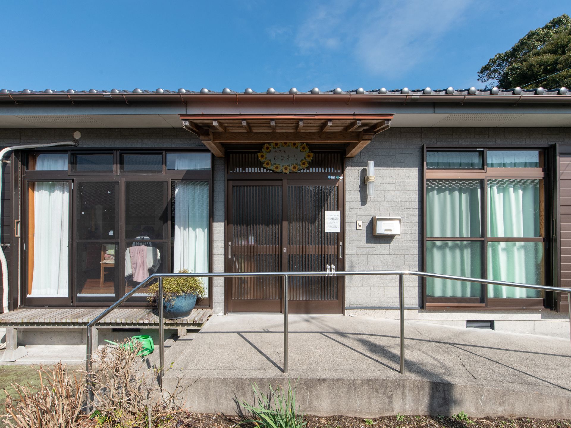 玄関前には手すりが設置された、緩やかなスロープがある。引き戸の玄関扉があり、外灯と郵便受けが設置されている。