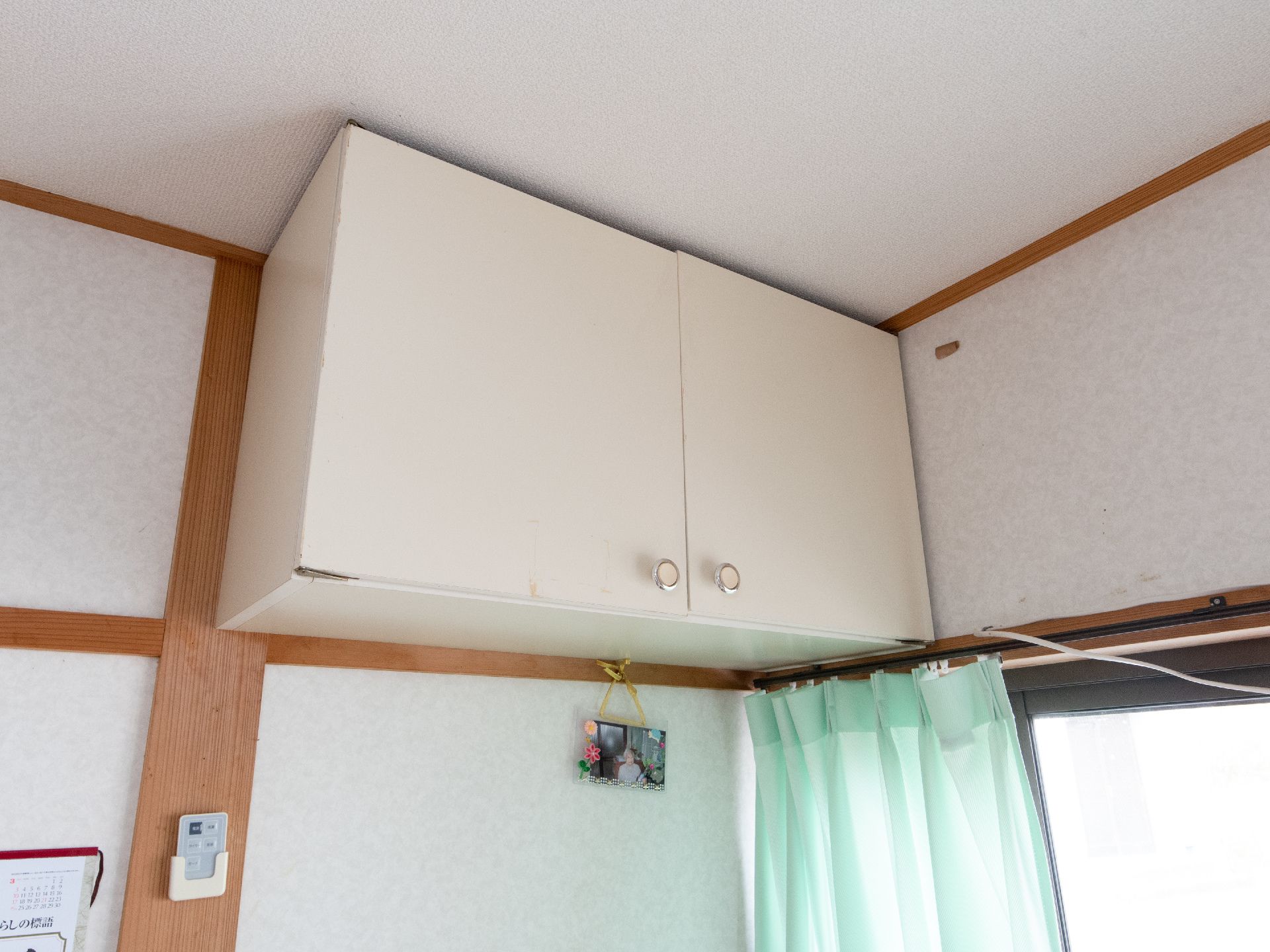 白を基調とした居室である。天井から吊り戸棚が取りつけられており、その下に写真が飾られている。また、緑のカーテンがかけられている。