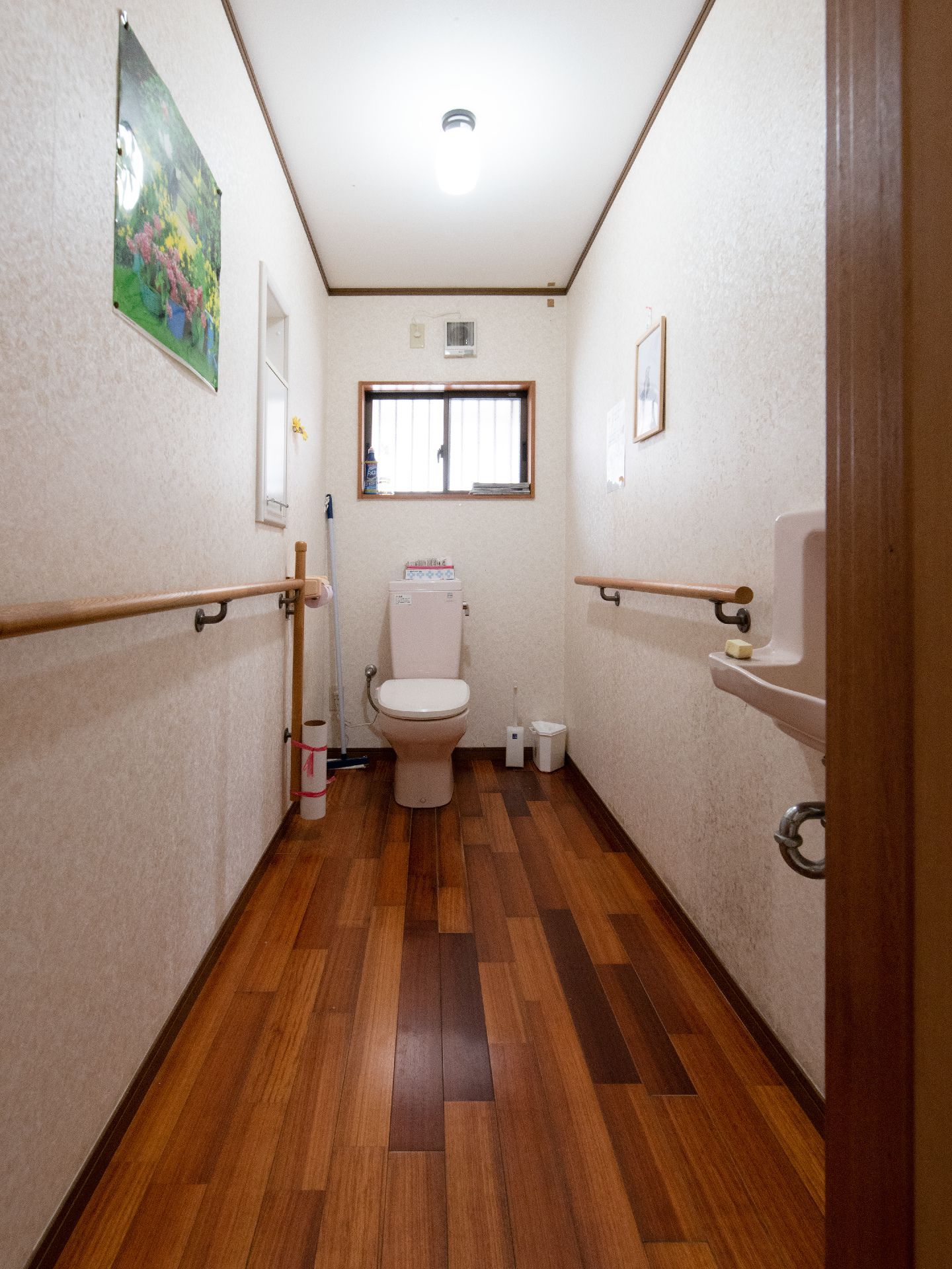 フローリングのトイレである。壁には絵が飾られており、奥には窓がある。また、入口には手洗い場も設置されている。