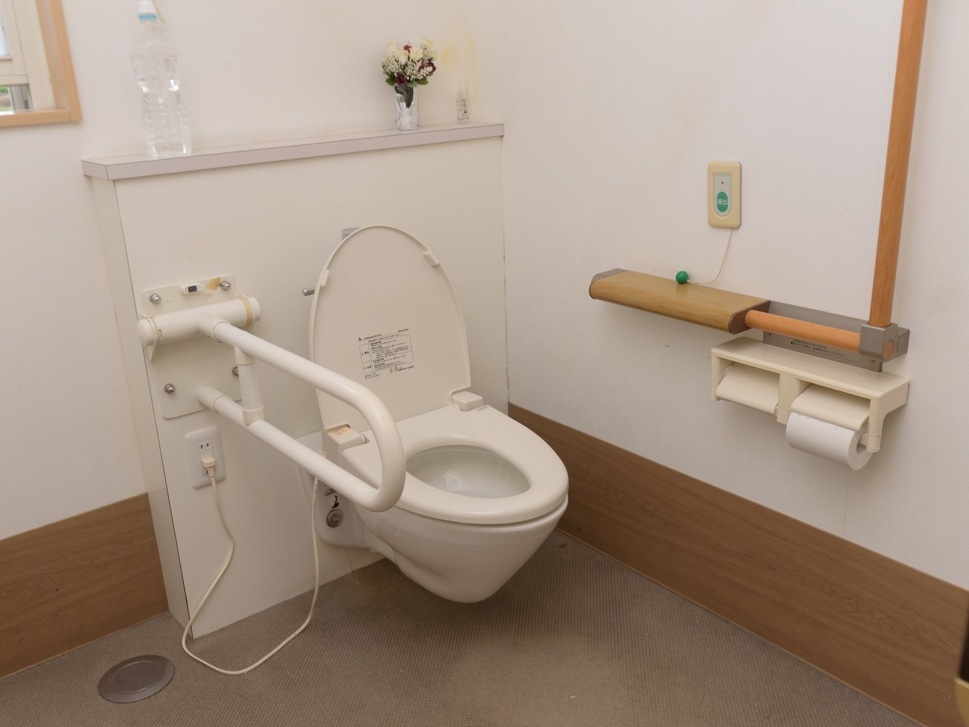 白い壁で囲まれたトイレで、壁に便座が直接取り付けられている。便座の横には折り畳み式の手すりがある。