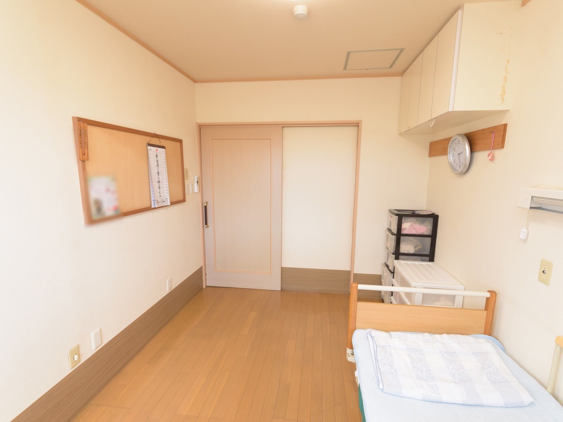 フローリング材を使用した居室で、奥に引き戸の入口がある。右手前に木製のベッドがあり、布団が敷かれている。