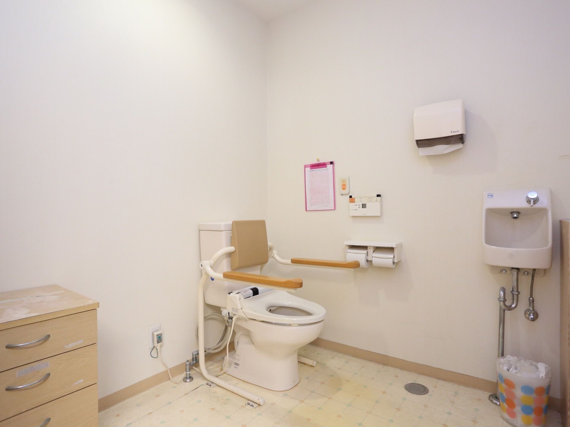 トイレ内の様子。洋式便器に背もたれ付の手すりが設置されている。壁に操作ボタンとナースコールが付いている。