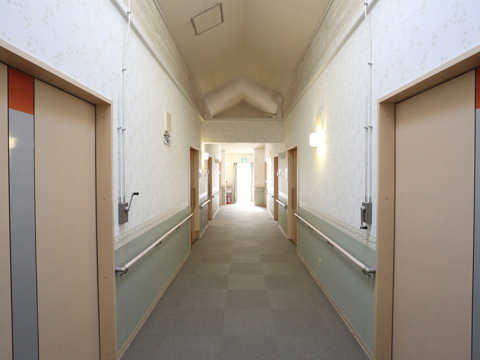 施設内の廊下の様子。両壁にはおしゃれな壁紙が貼られ、手すりが設置されている。床にはタイルカーペットが敷かれている。