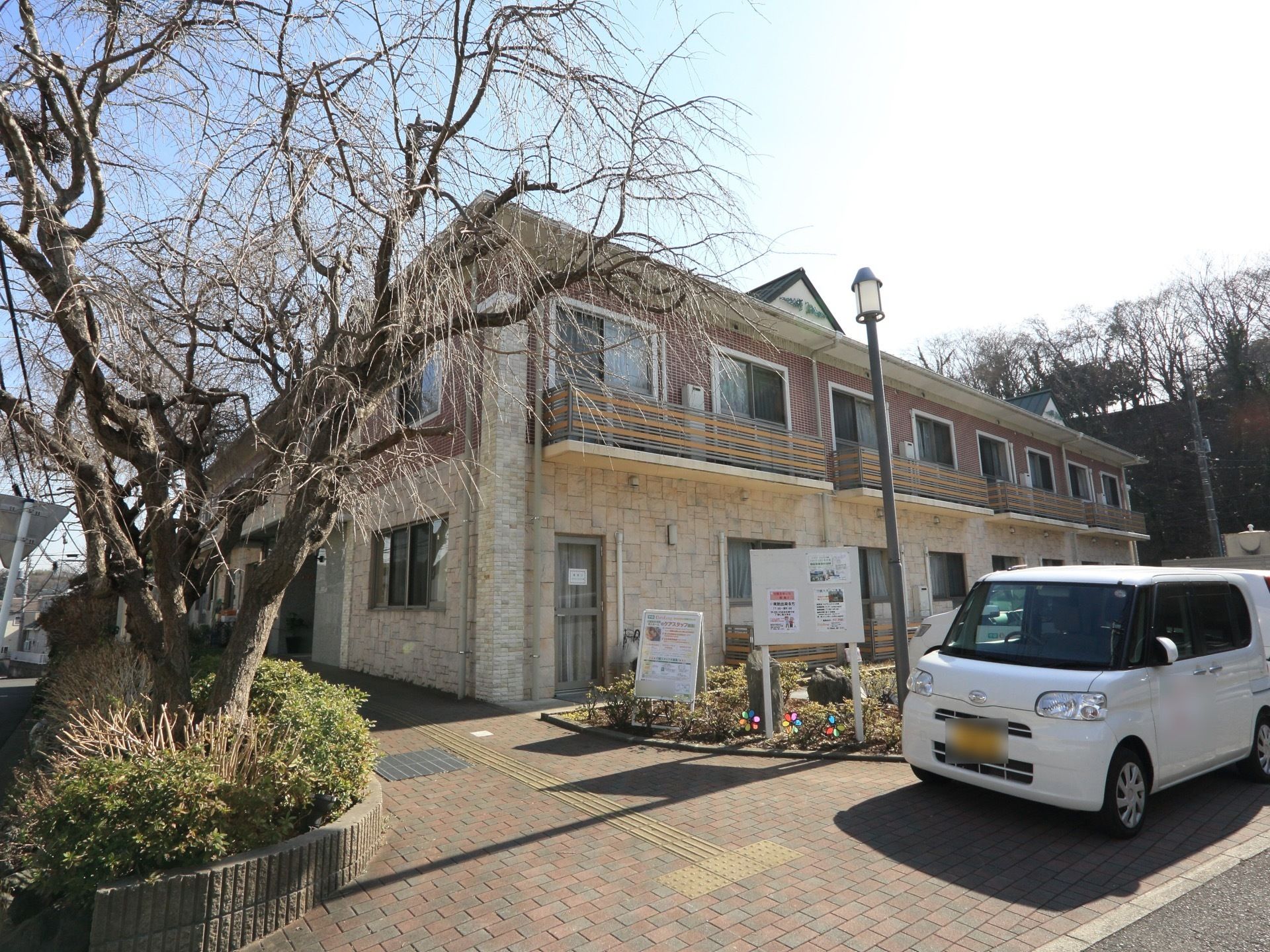 ２階建ての施設は、ブラウン系の色でまとめられた落ち着いたデザインである。建物の周辺には植込みがあり、左端に大きな木がある。