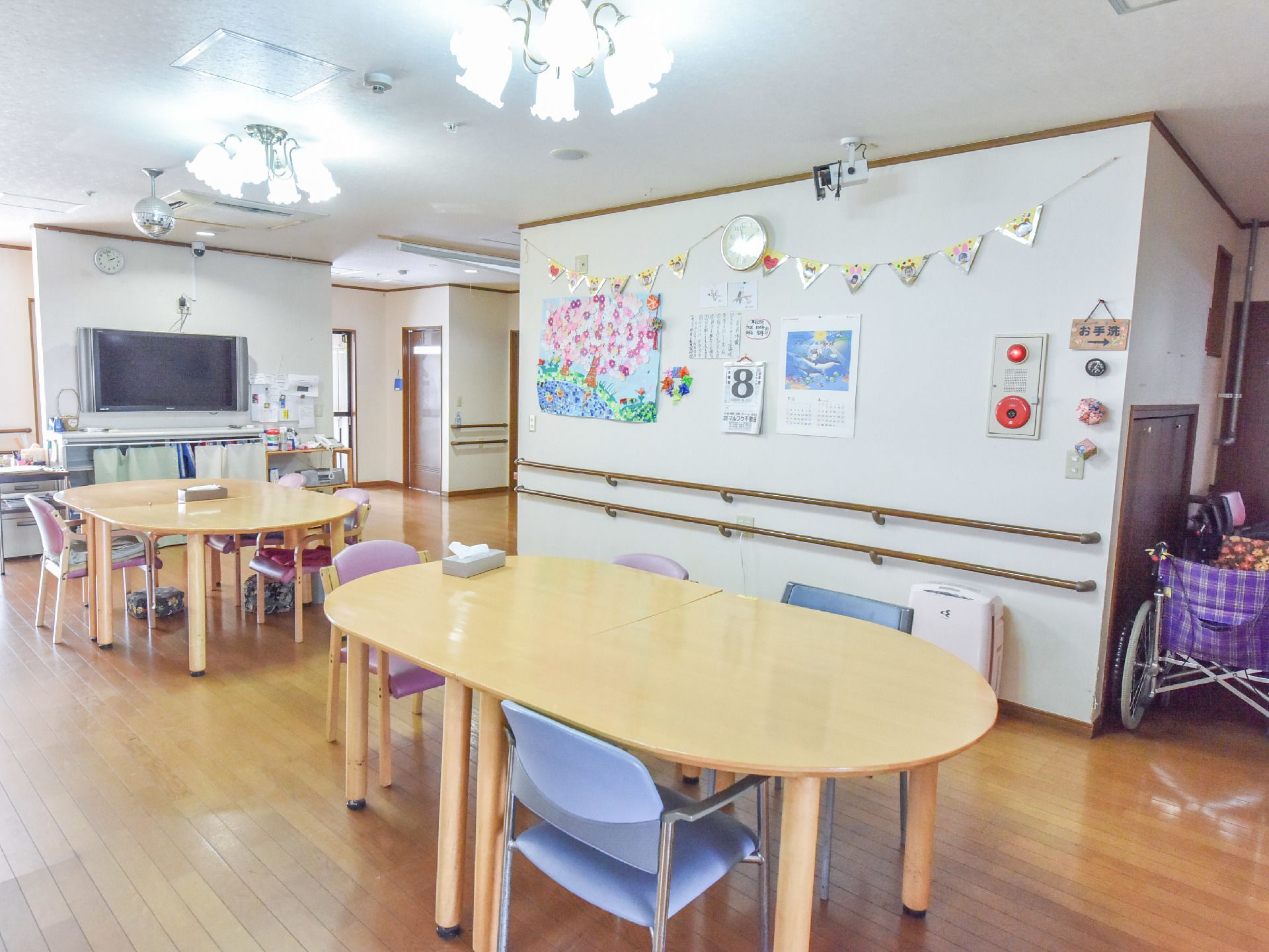 楕円型のテーブルと椅子が配置されている。壁には手すりがある。大型テレビが設置されている。火災報知器がある。