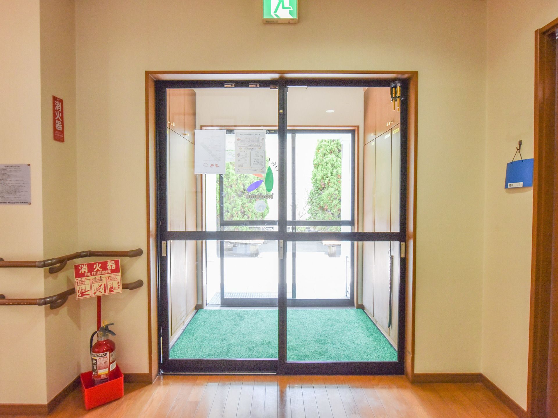風除室に下駄箱がある。入り口ドアは引き戸式である。消火器が配置されている。壁には手すりがある。非常口表示がある。