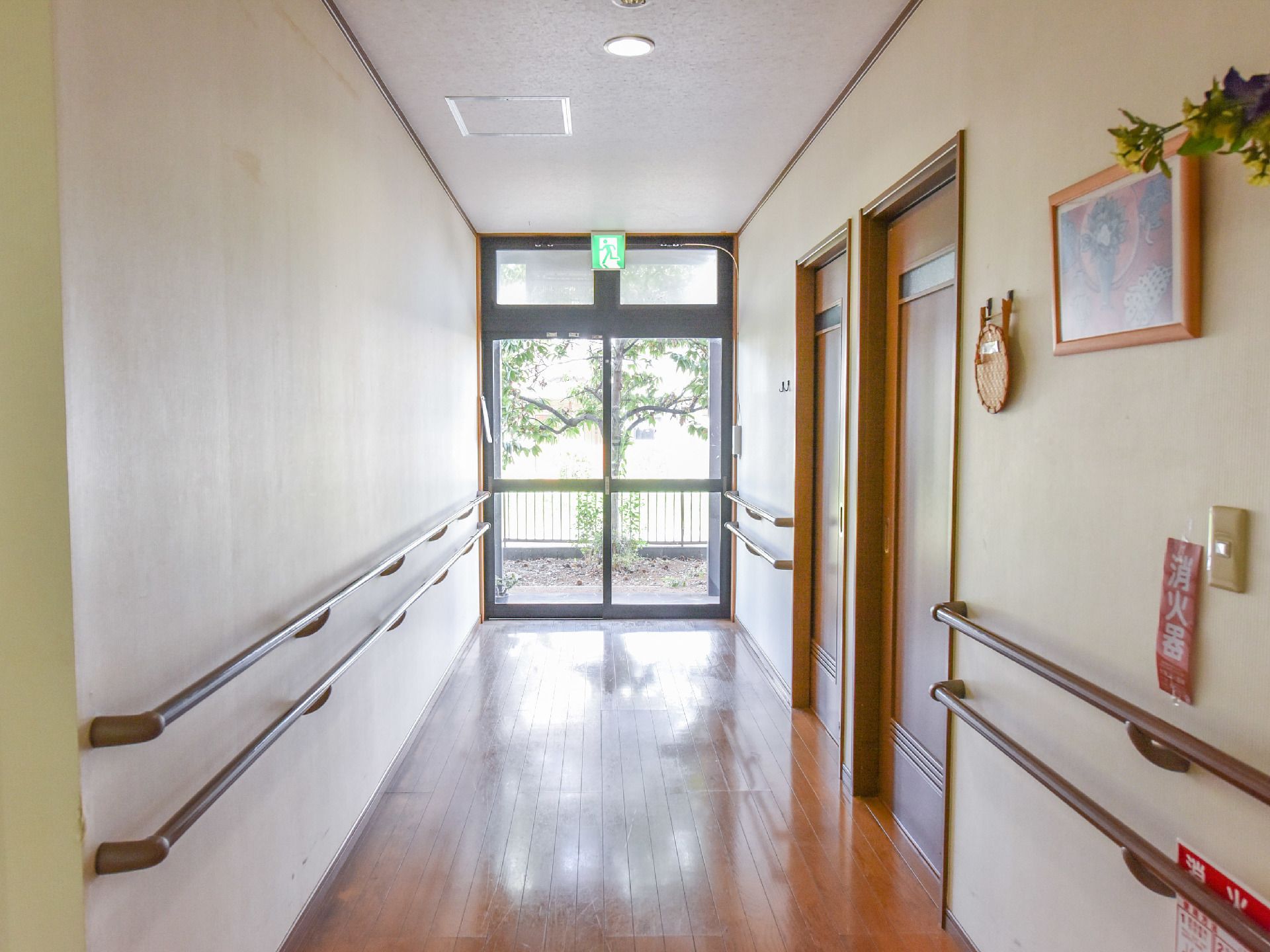 通路両脇に手すりが設置してある。消火器がある。非常口表示がある。通路奥の窓から明るい陽射しが差し込んでいる。