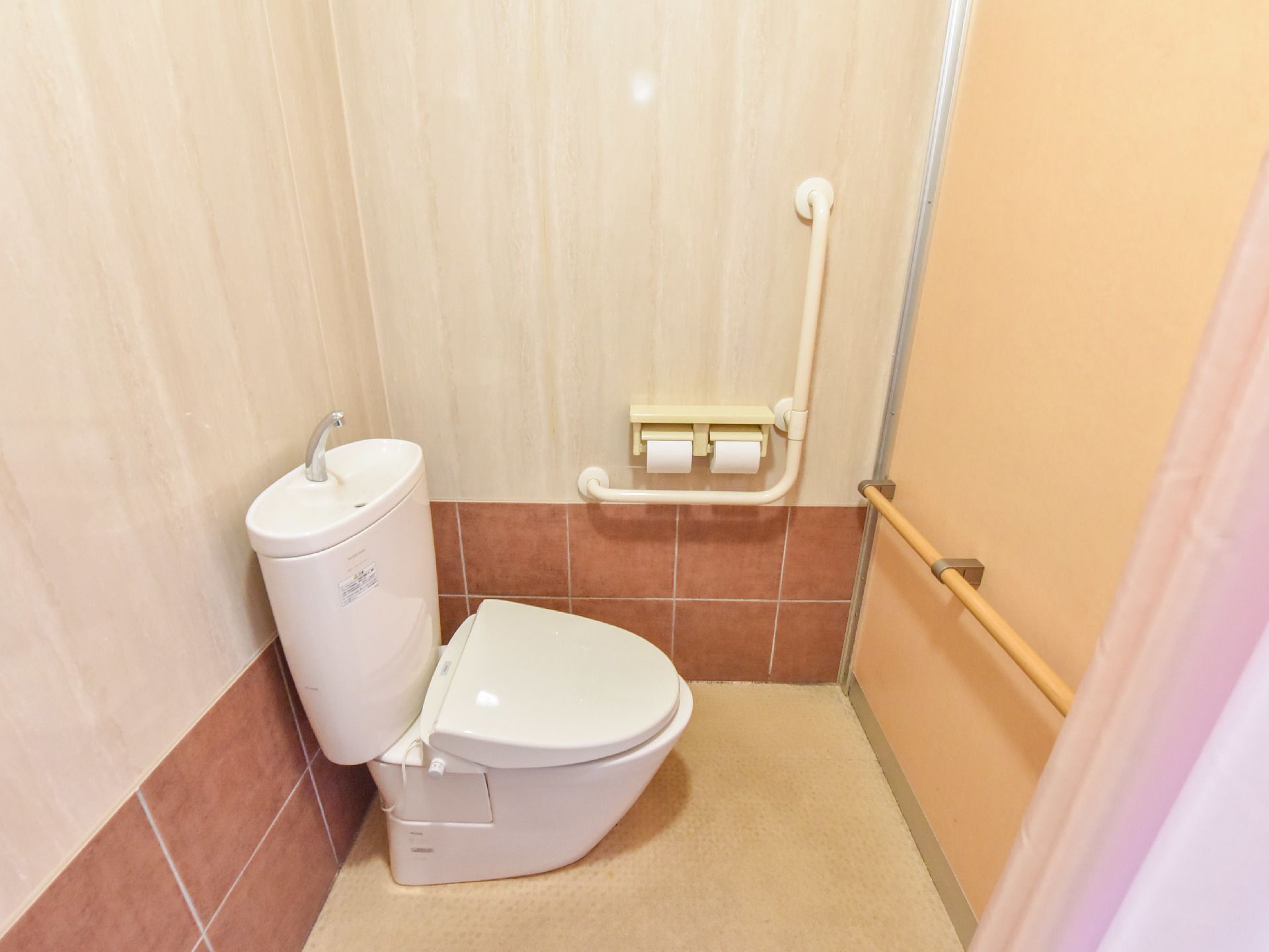 壁に2か所手すりが設置してある。洋式トイレで、手洗い用のシンクがある。仕切りにはカーテンが用いられている。