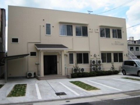 雲がかかる青空の下に、白い２階建ての建物が建っている。建物の前にエアコンの室外機が置いてあり、コンクリートで整備された駐車場がある。