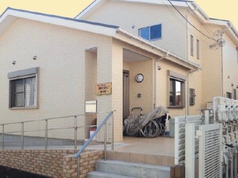 ベージュ色で統一された柔らかい雰囲気の2階建ての施設外観。玄関まで緩やかなスロープと数段の階段があり、どちらにも手すりが設置されている。
