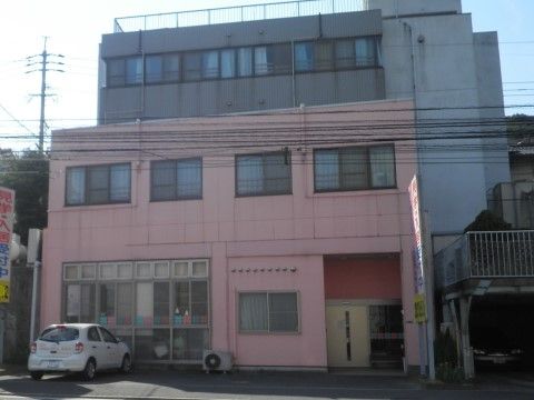 ピンク色の外壁が特徴的な建物である。１階には背の高い窓が設置されており、近くにエアコンの室外機が置かれている。