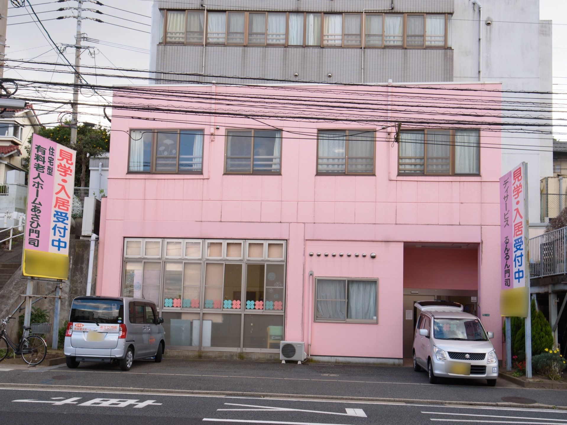施設の外観はピンク色で、窓には青とピンクの花の飾りが貼られている。施設の前に数台の駐車スペースがある。