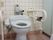 施設のトイレはタイル壁に茶色の床、薄青のウォシュレット水栓トイレ。簡単に水を流せるタイプで壁には可動式の手すりが設置されている。