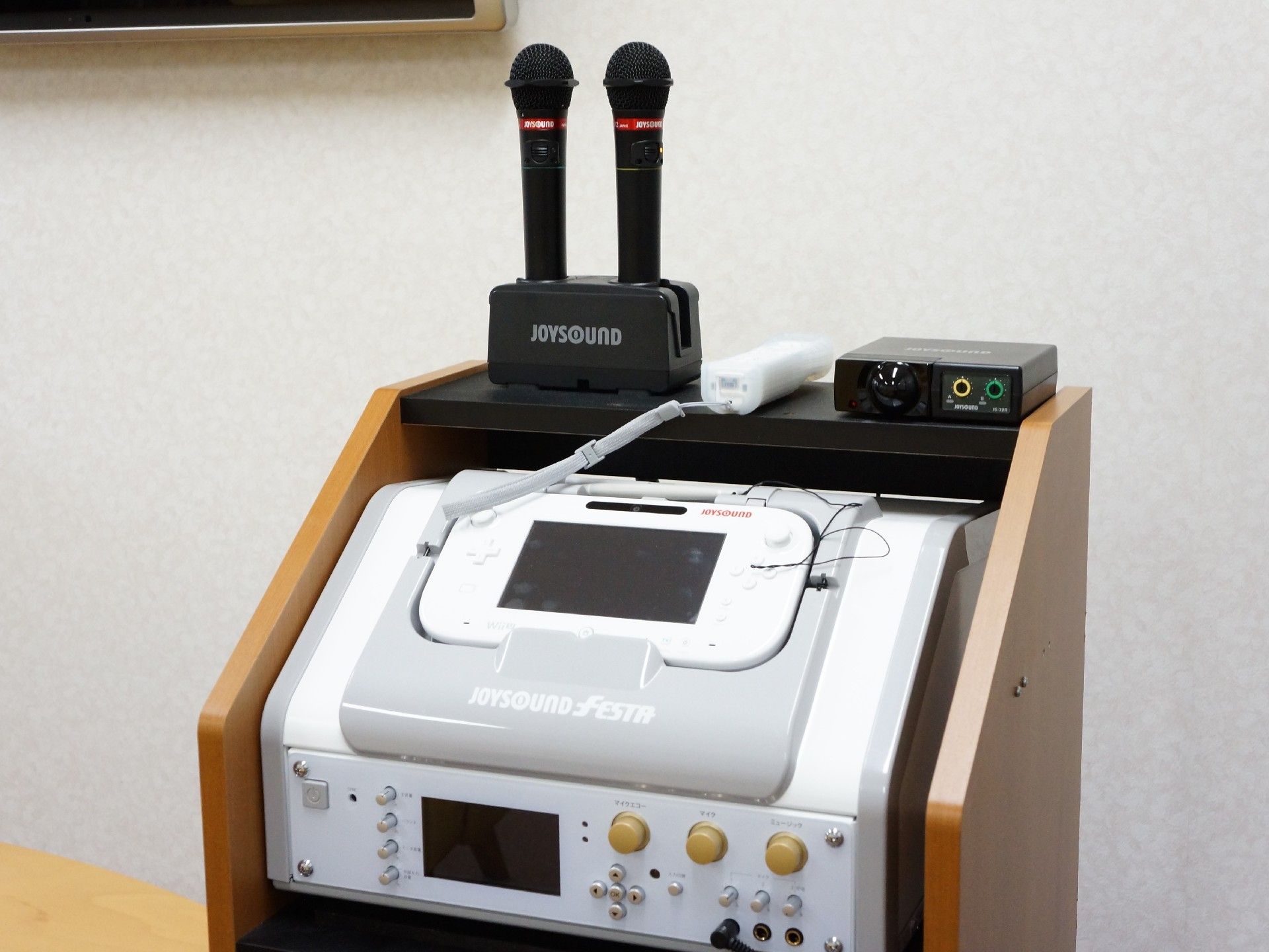 カラオケの機材のアップが写されている。機材はジョイサウンド製のフェスタが設置されている。リモコンがありマイクが2つ置かれている。