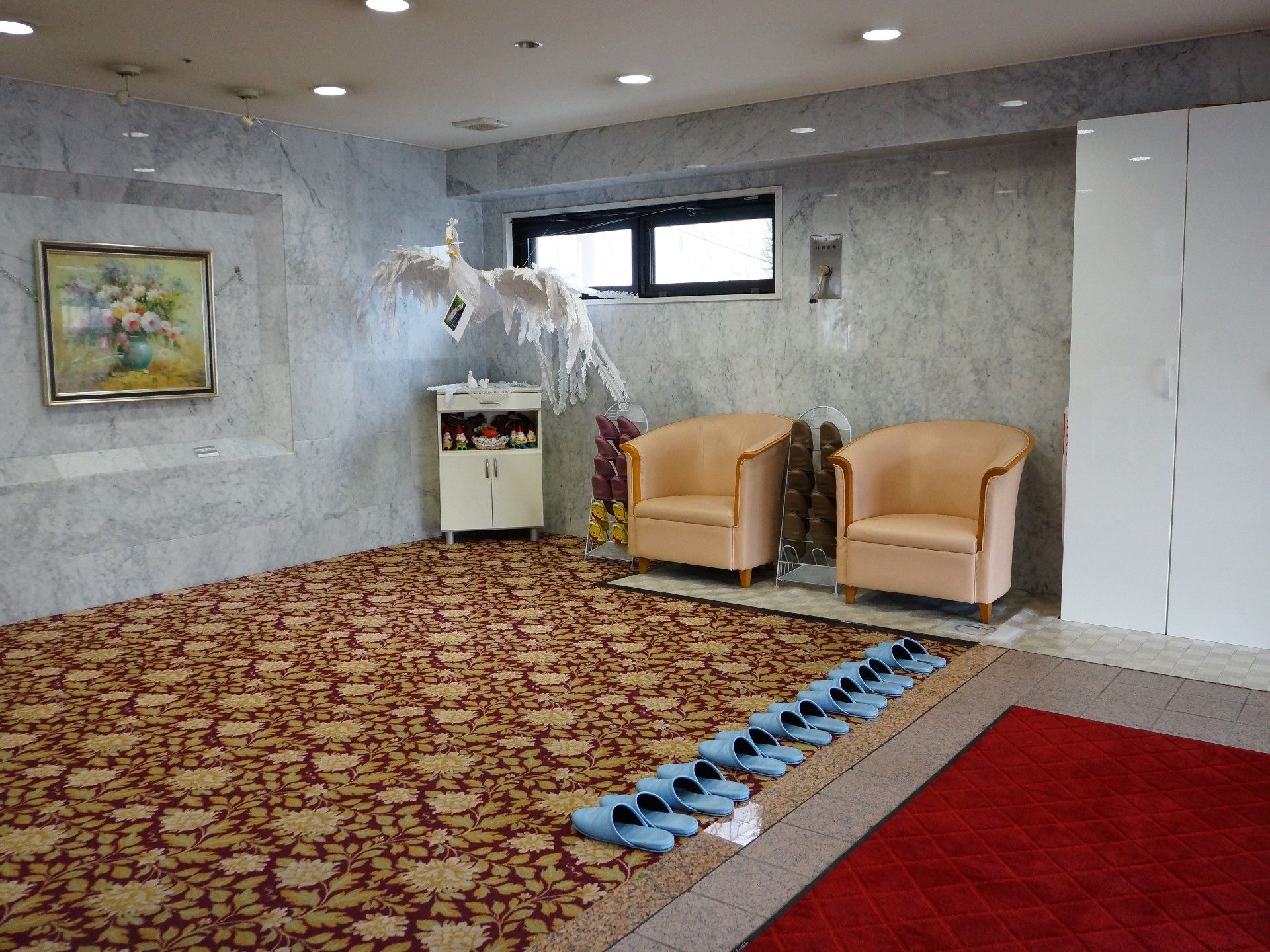施設のエントランスには絨毯が敷かれ大理石調の壁には絵画があり椅子が2つ置かれている。棚もあり鳥の置き物も置かれている。