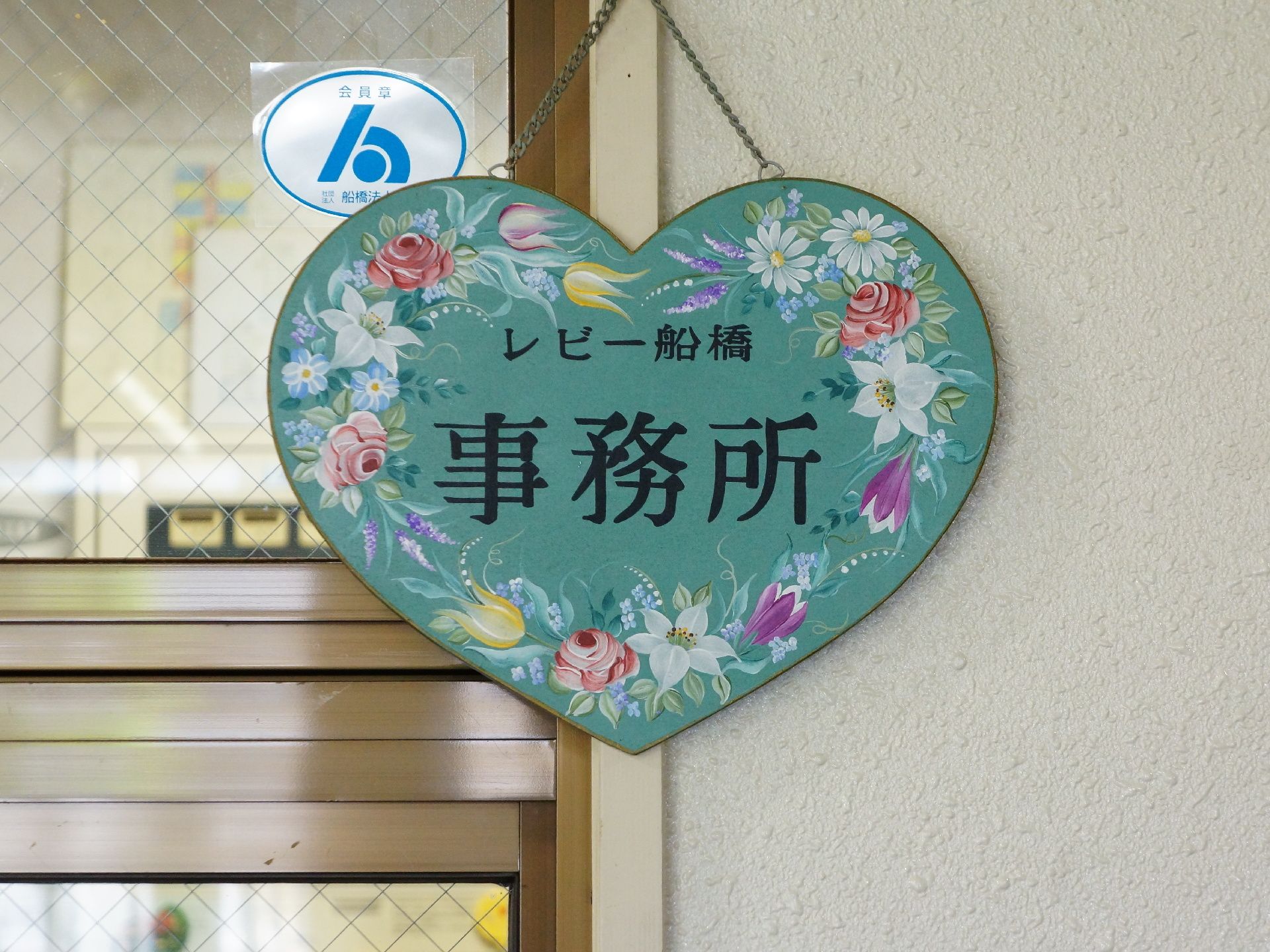 事務所の受付に掲げられた案内表示。ハートの形をしており、緑の板の上に色とりどりの花が美しく描かれている。