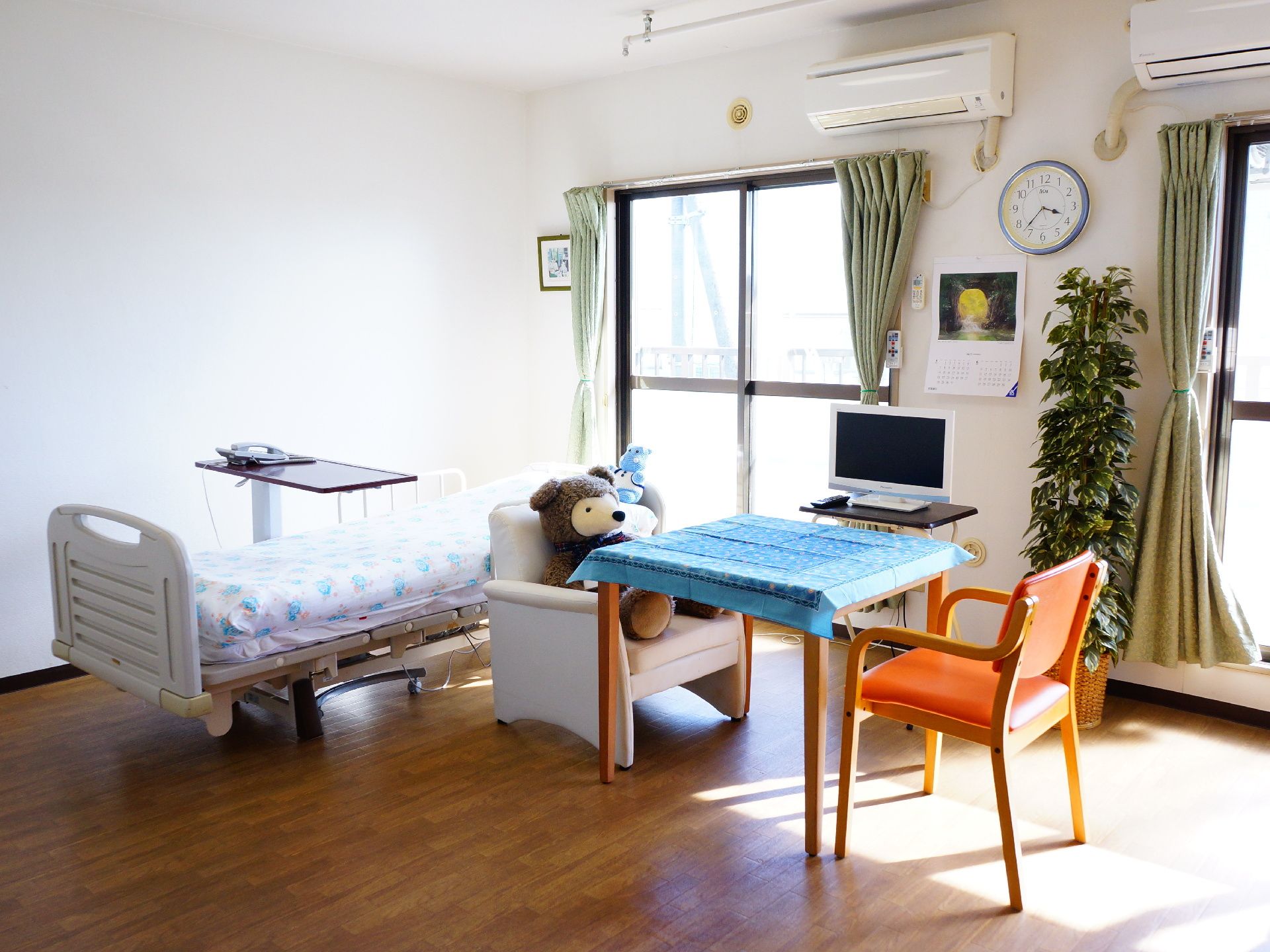 上下いっぱいに窓がある明るい部屋にベッドが設置してある。そばにテーブルとイスがあり、イスの上にはクマのぬいぐるみが置かれている。